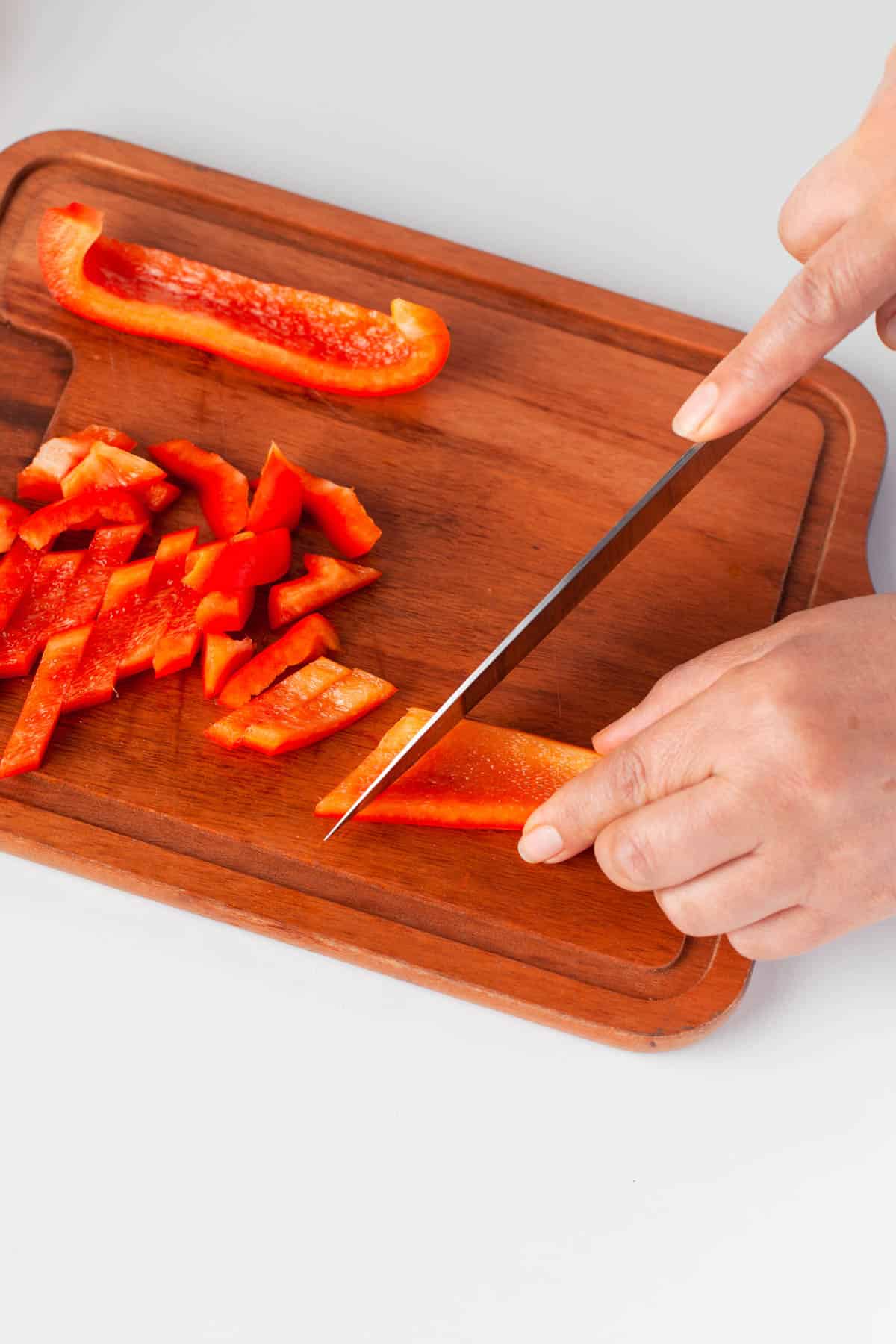 A hand slicing red bell pepper with a knife.