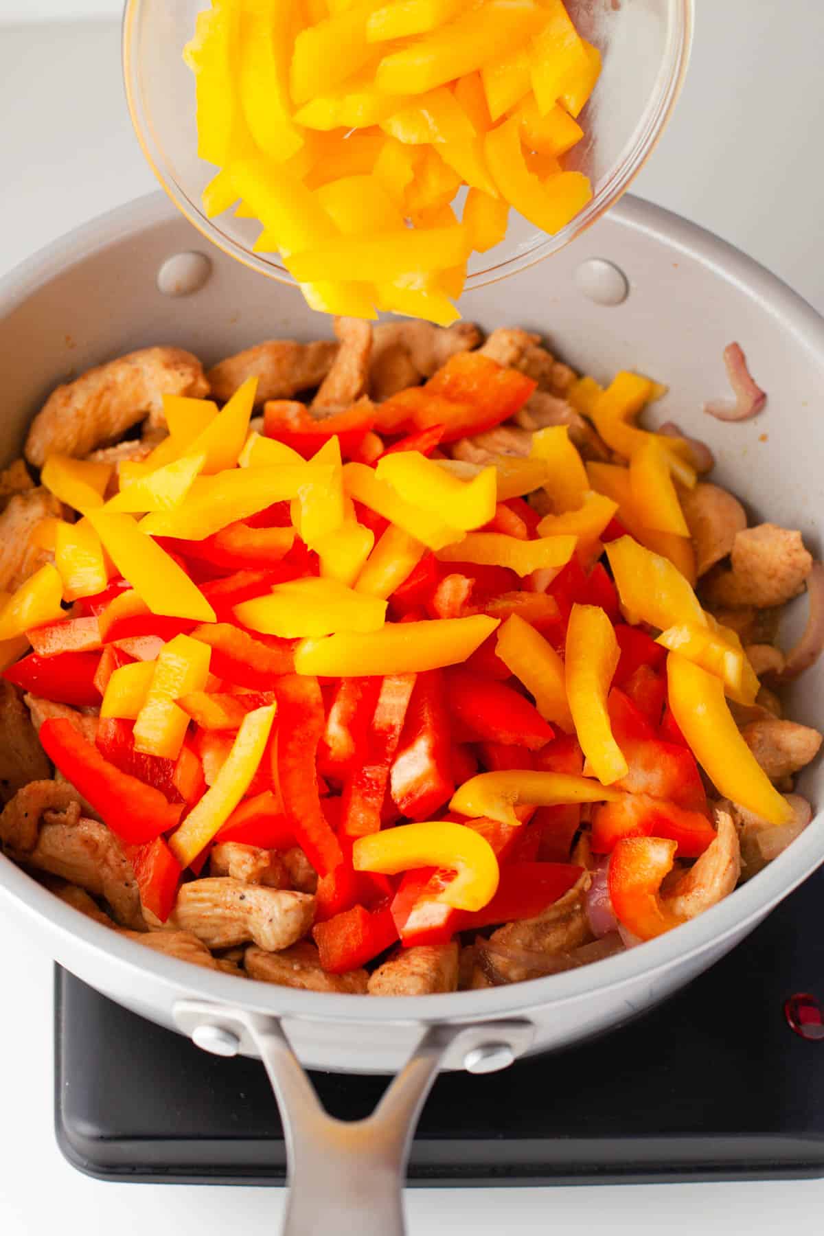 Adding sliced red and yellow bell peppers to cooked fajita chicken in a pan.