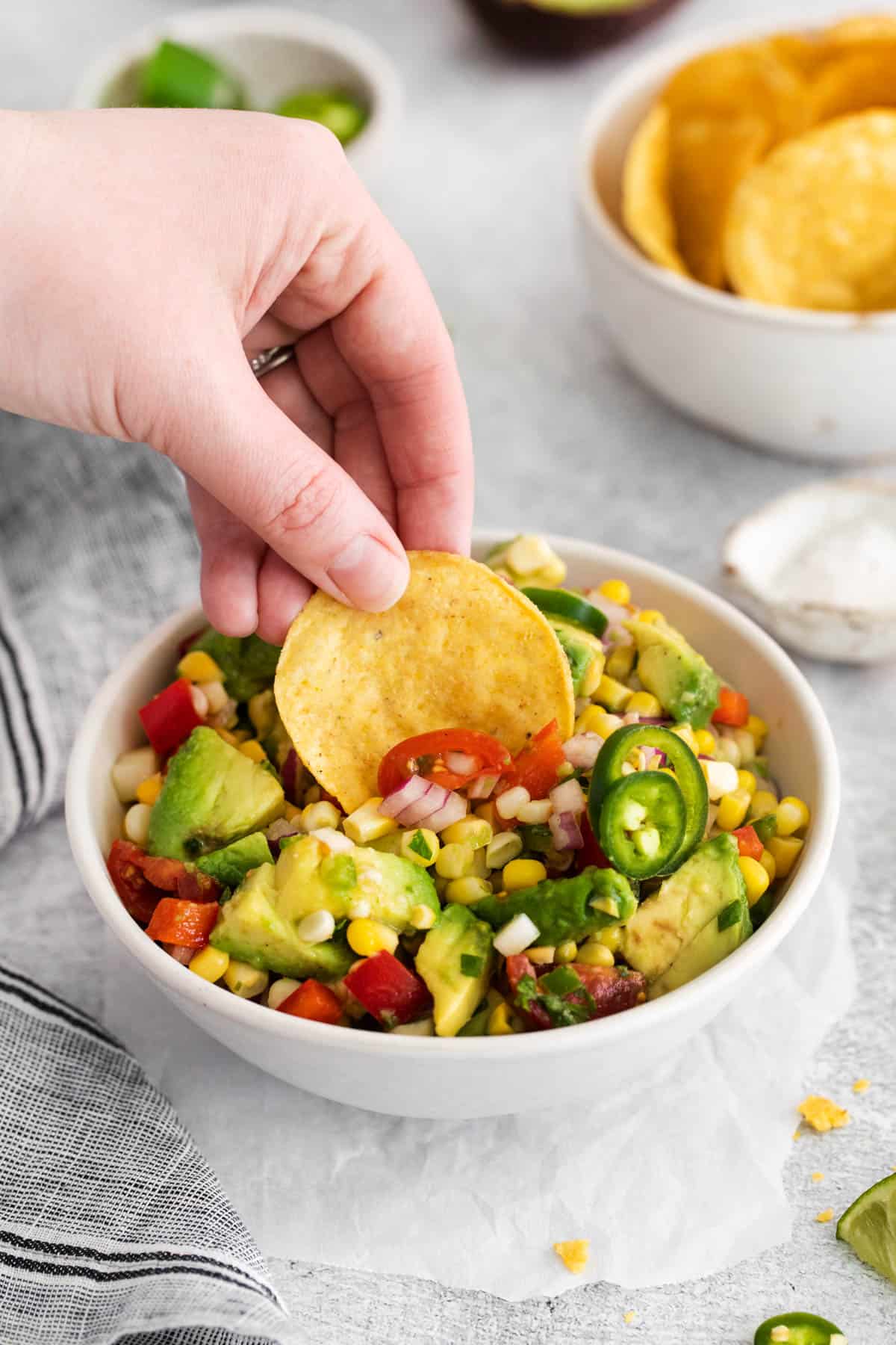 A hand holding a chip to scoop corn & avocado salad from a bowl.