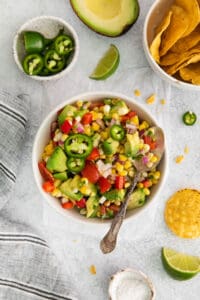 An overhead image of a bowl of corn and avocado salad with a spoon in it next to jalapenos, avocados, limes, and tortilla chips.