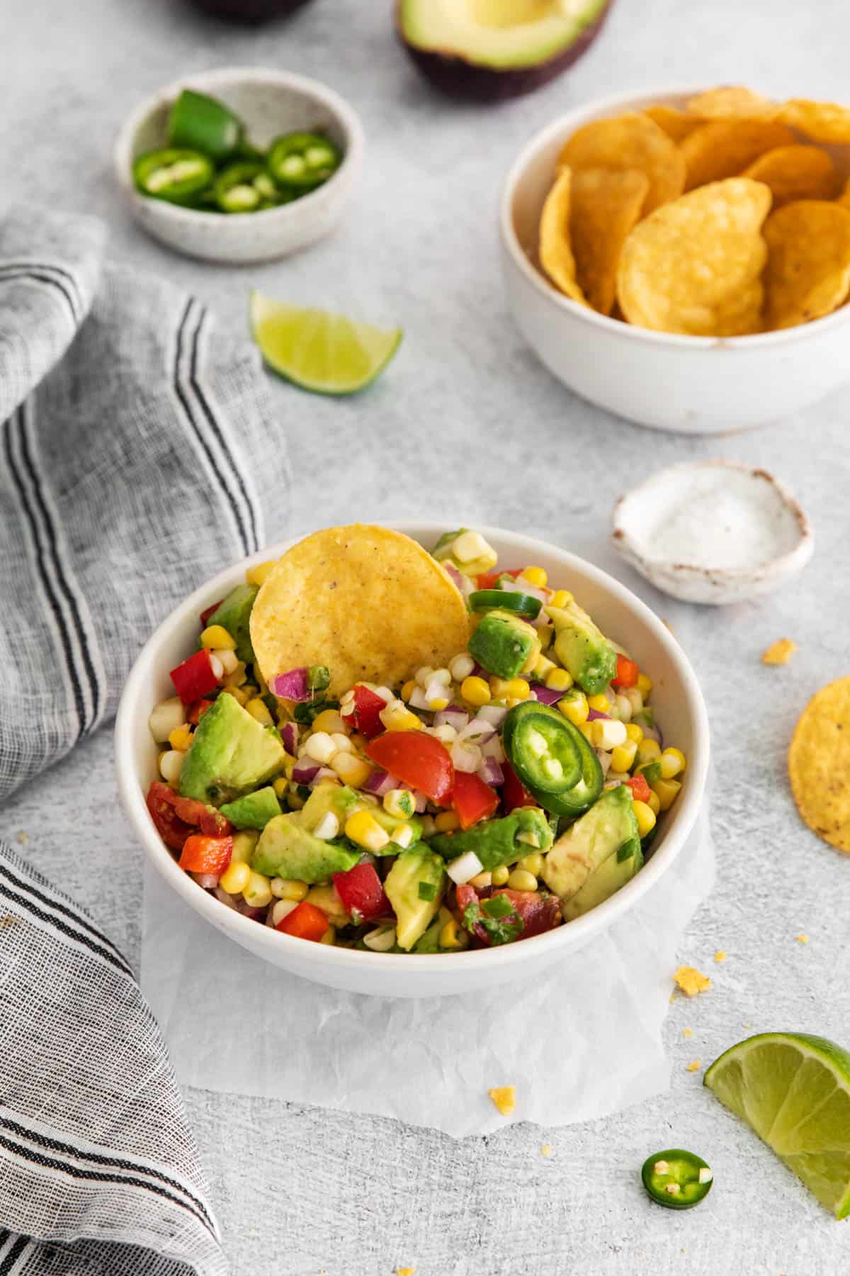 A bowl of corn and avocado salad in front of bowls of corn tortilla chips, limes, and sliced jalapenos.