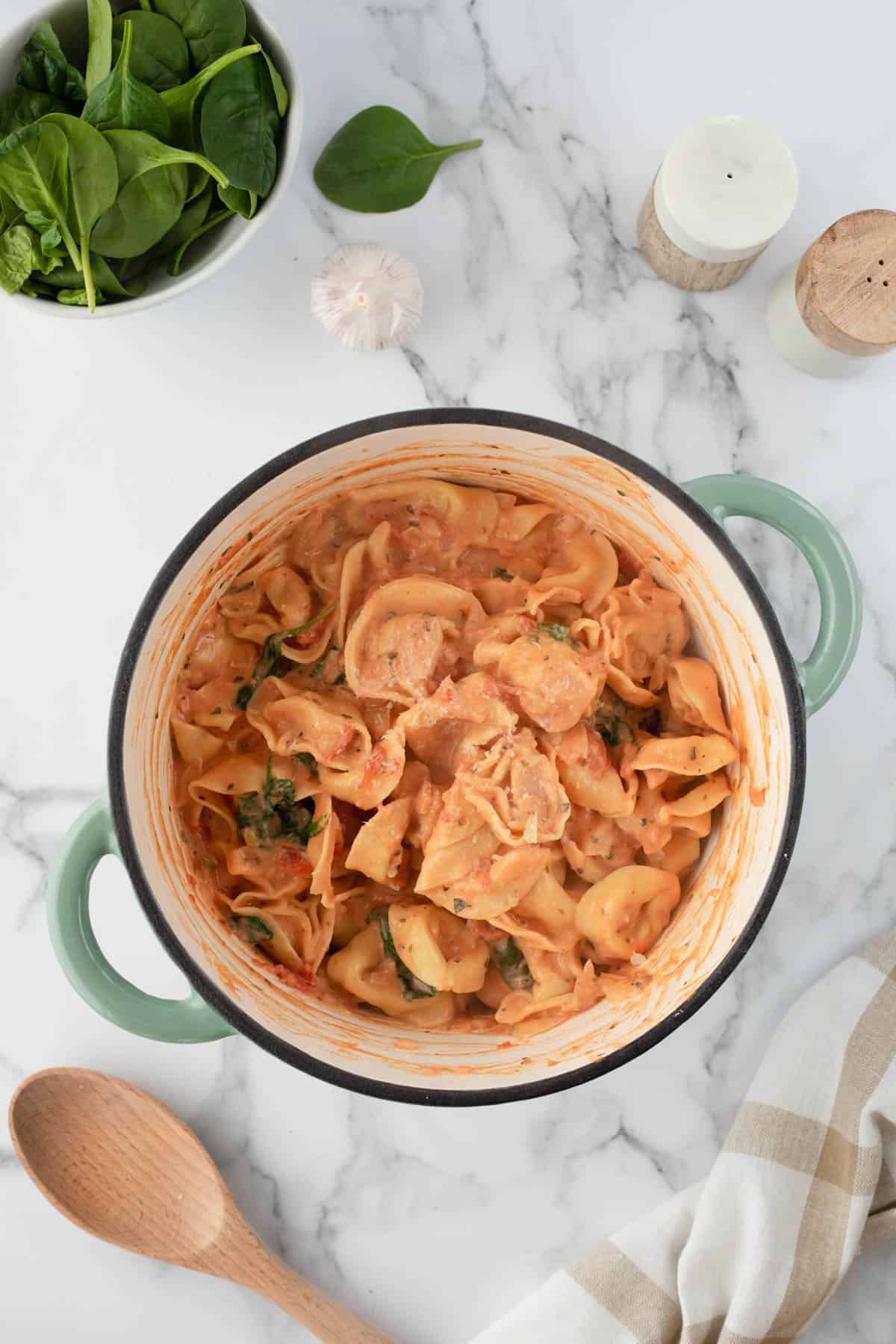 A pot of creamy tomato and spinach tortellini next to a bowl of spinach and a wooden spoon.