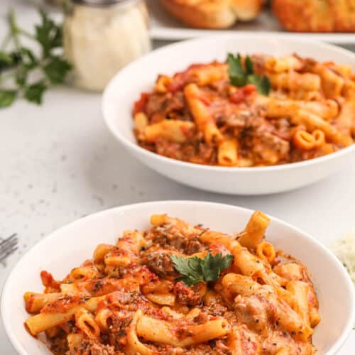 Bowls of crock pot baked ziti pasta in front of a plate of garlic bread.