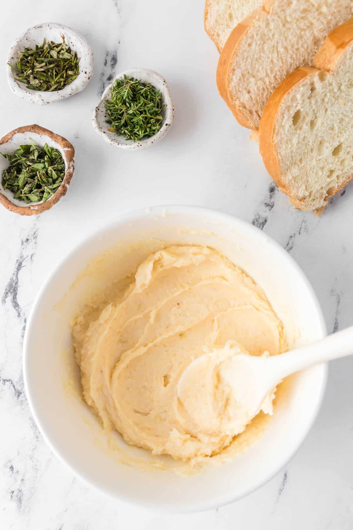 A spoon being used to mix softened butter in a white bowl next to small bowls of herbs and sliced Italian bread.