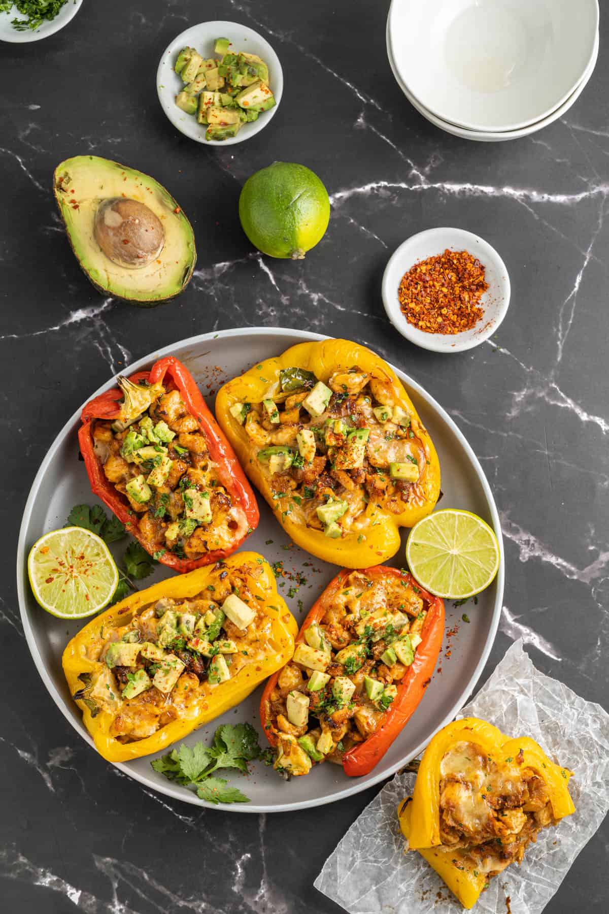 A plate of fajita chicken stuffed peppers on a black surface with avocado, lime, and spices around them.