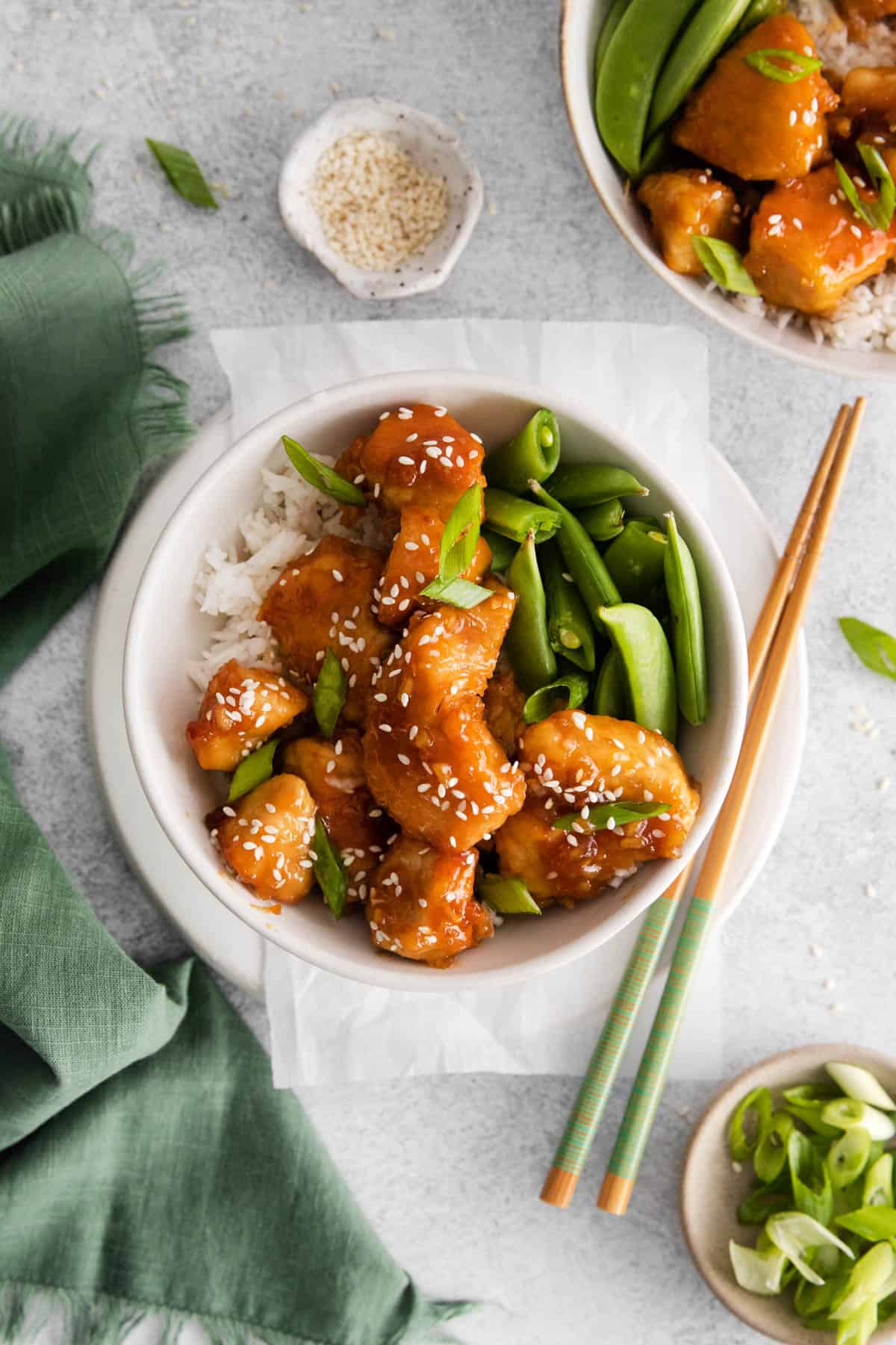 An overhead image of sticky honey garlic chicken with sesame seeds sprinkled on top next to bowls of sesame seeds, sugar snap peas, and more chicken.
