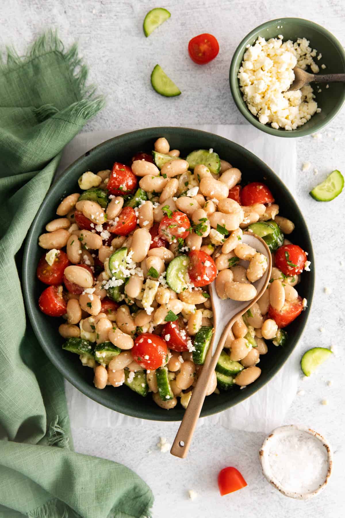 A spoon in Mediterranean white bean salad on a green plate next to a green cloth napkin surrounded by cherry tomatoes, cucumber slices, and feta cheese.