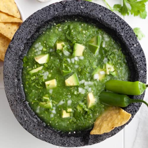 A close image of salsa verde in a bowl with avocado chunks, tortilla chips, and jalapenos on top for garnish.