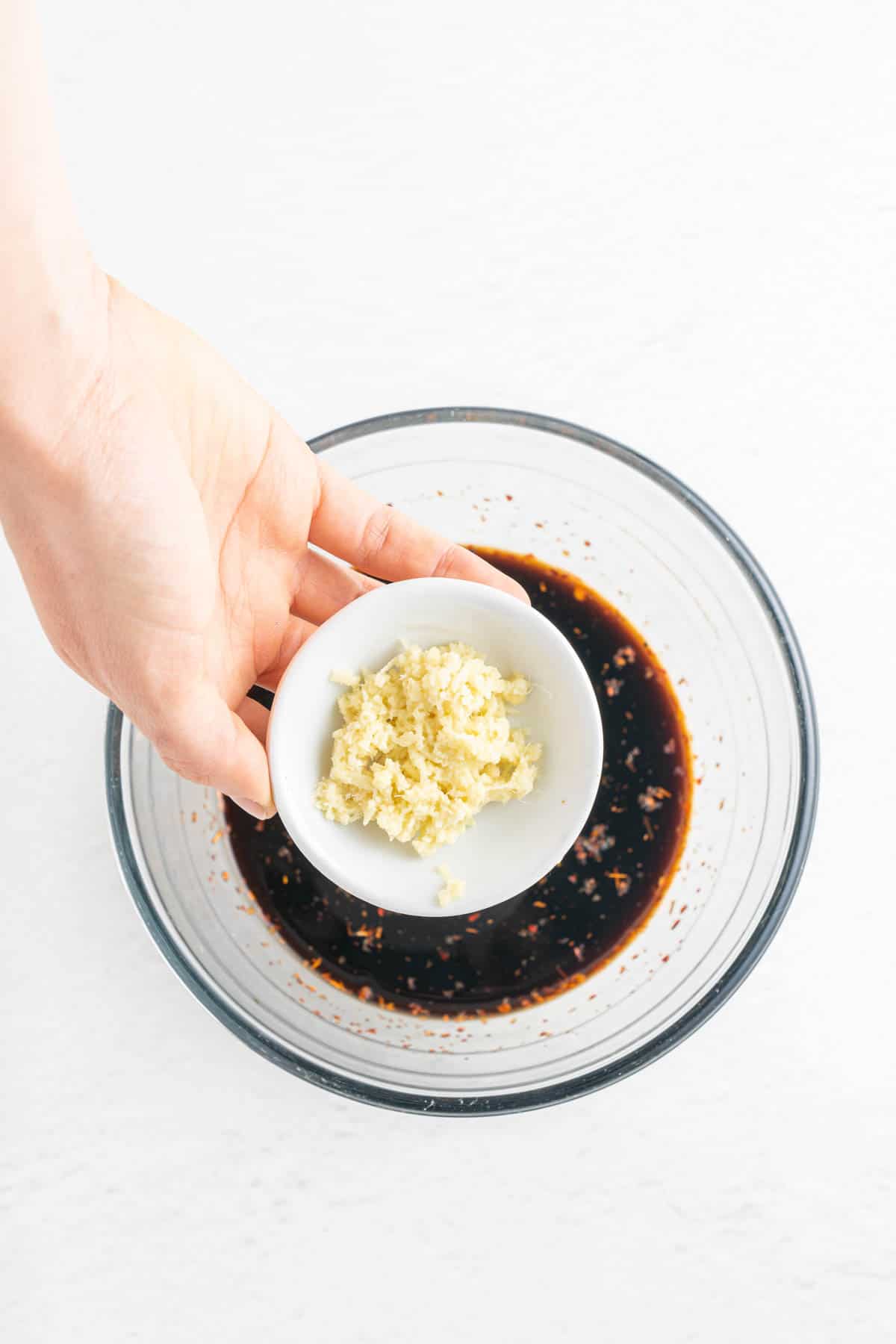 A hand holding a bowl of minced garlic over soy sauce and other ingredients.