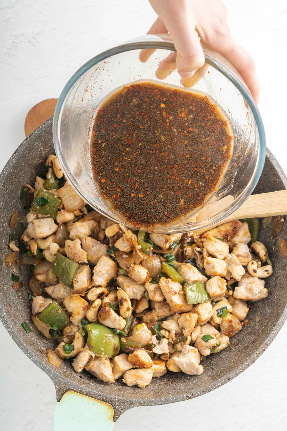 Adding sauce to chunks of cooked chicken, cashews, and vegetables in a pan.