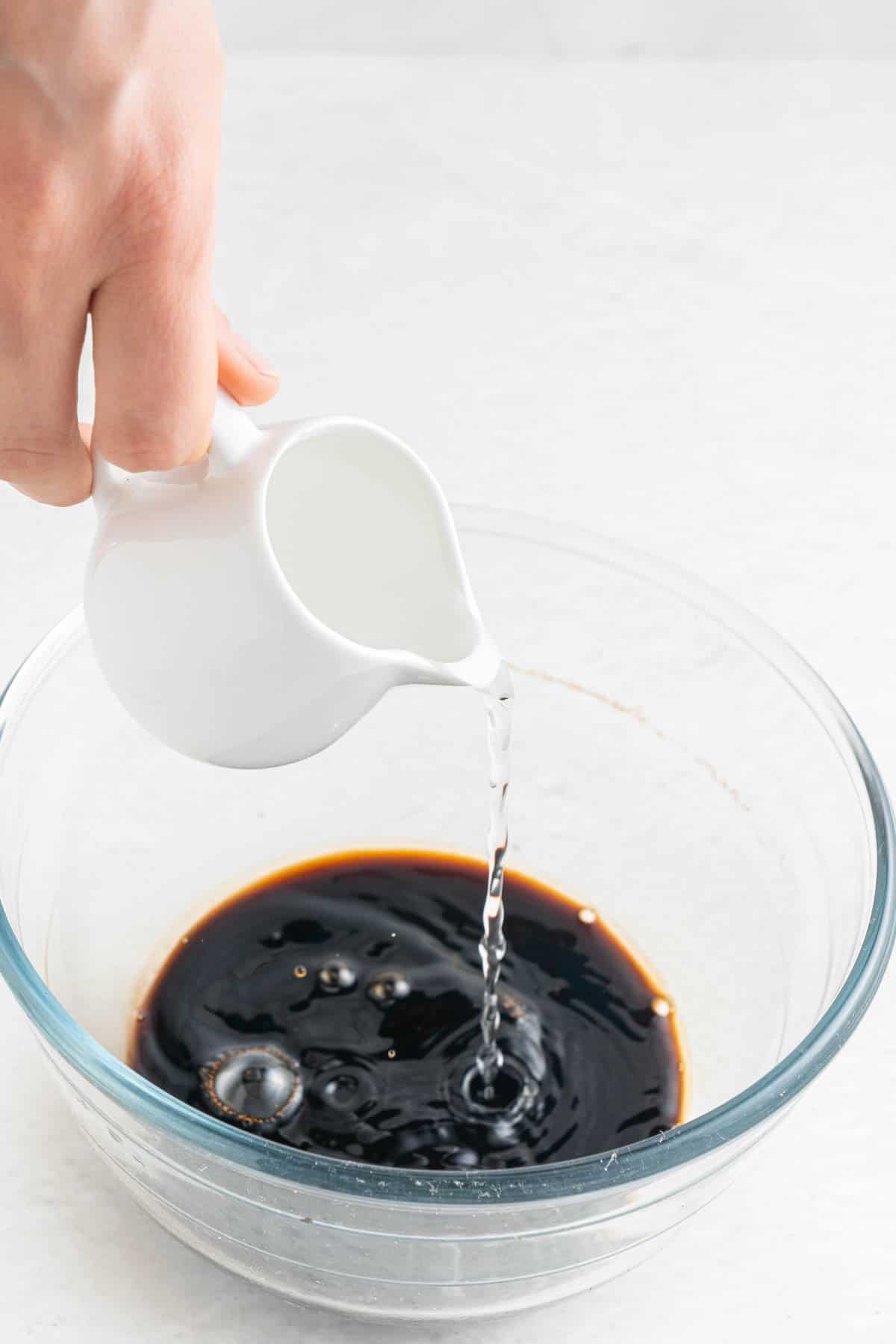 A small white pitcher pouring water into a glass bowl with soy sauce.