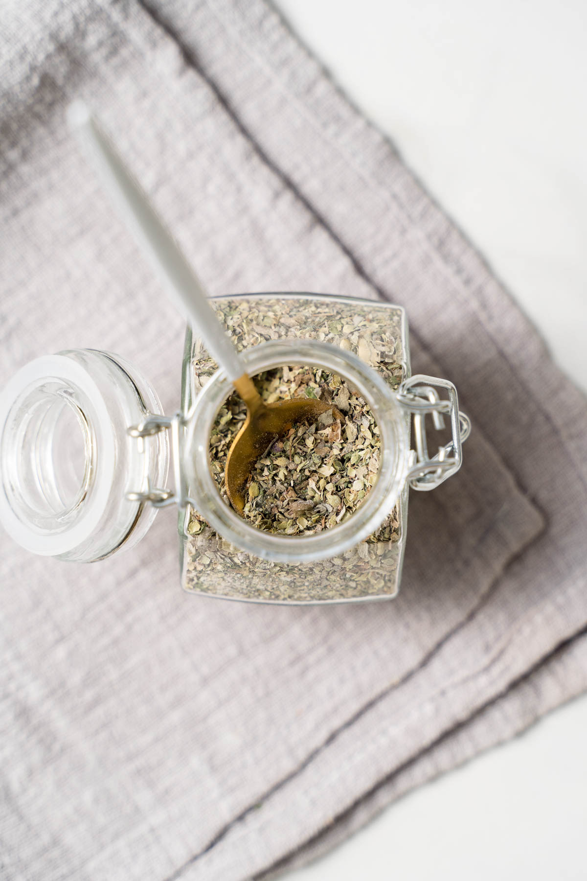 Image of greek seasoning in a glass lidded jar with a spoon resting in the spice blend. 