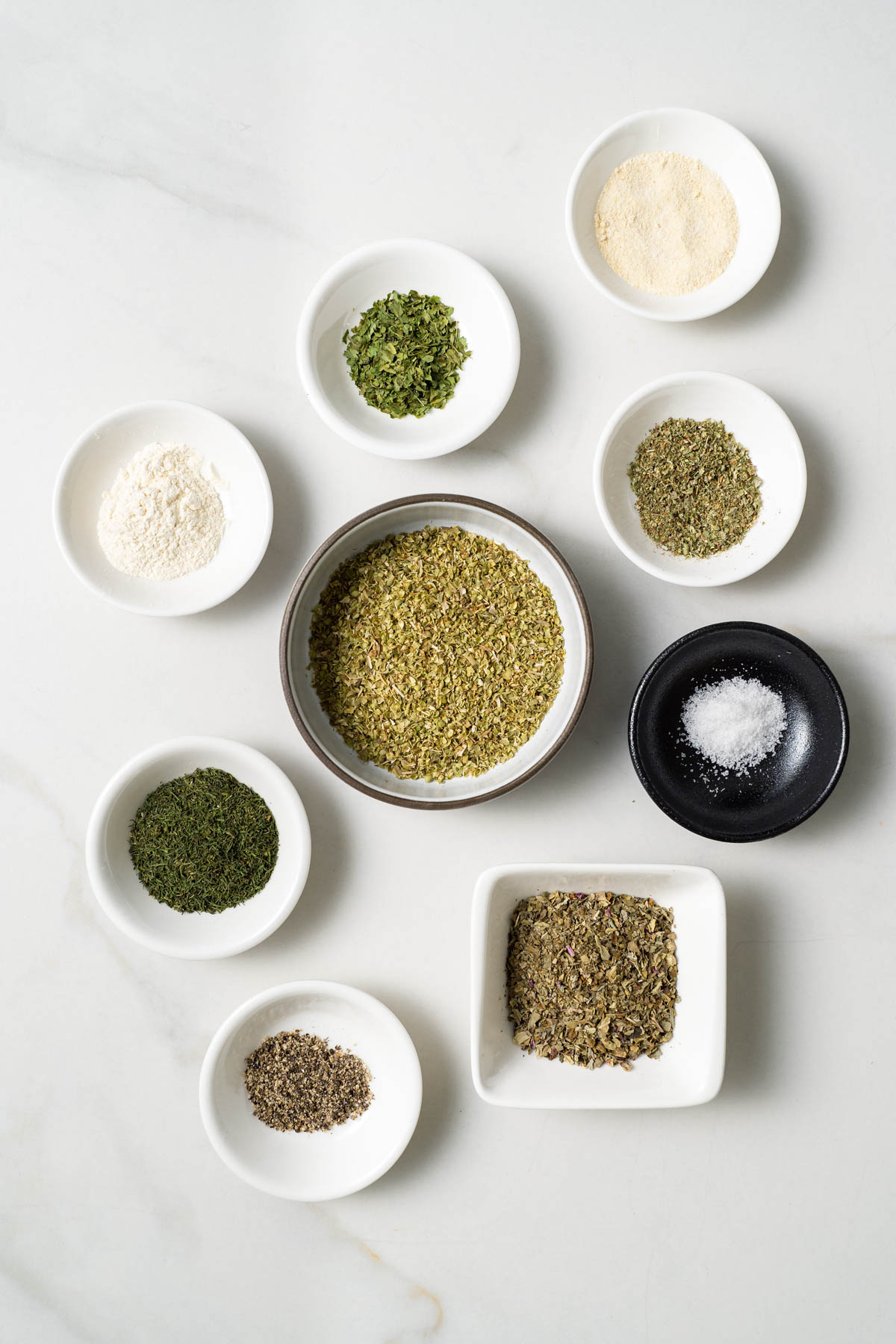 Individual bowls containing herbs and spices are displayed on a white surface. 
