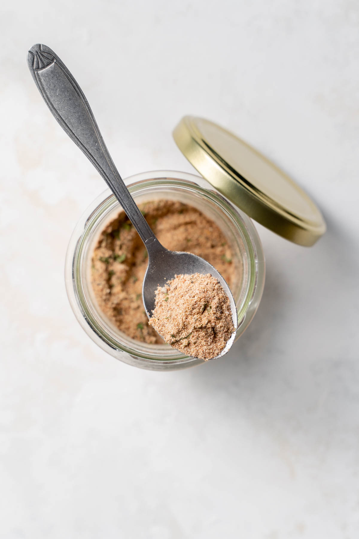 Overhead view of a jar of homemade jerk seasoning with a spoon holding some spice and a lid resting on the side of the jar.