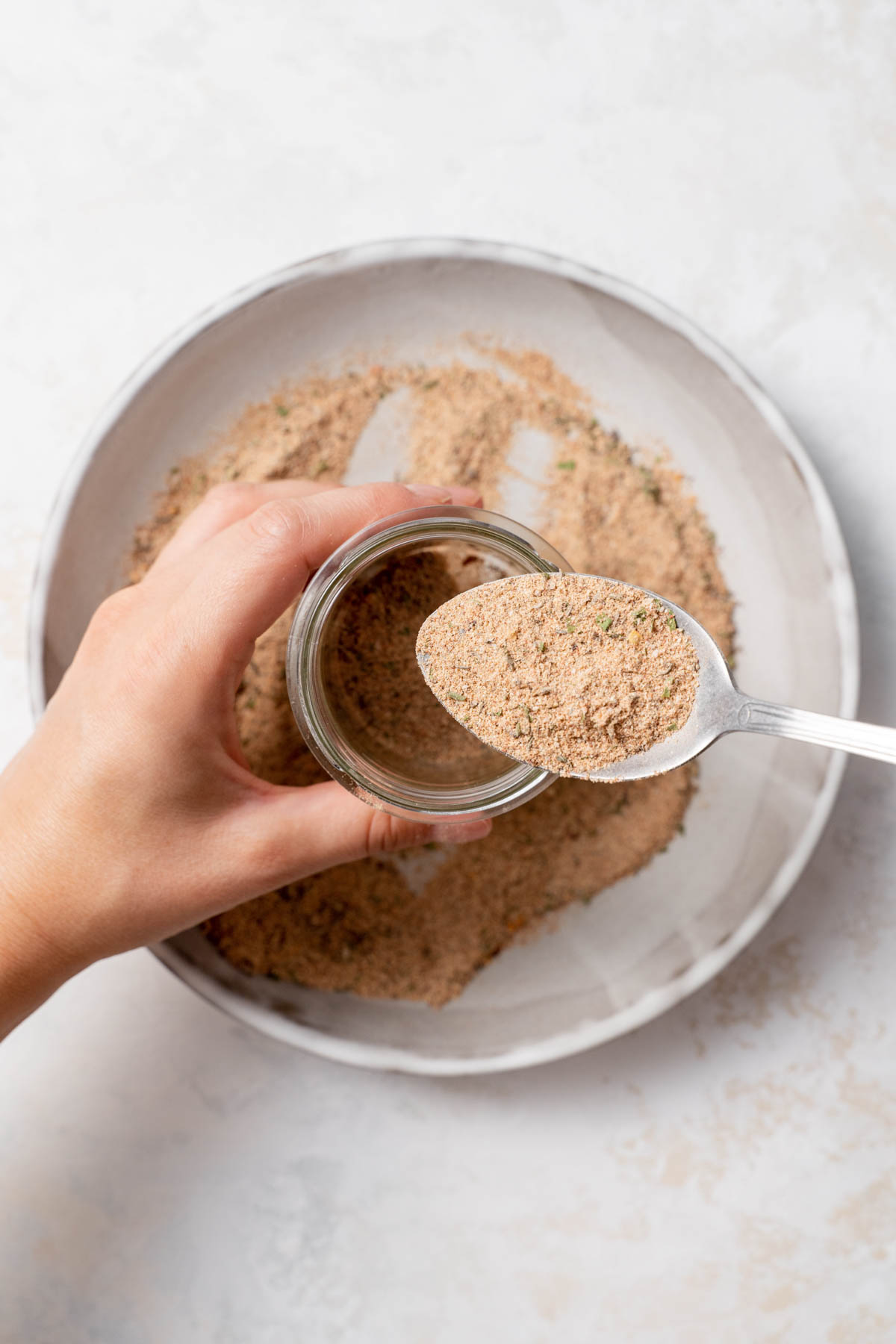 Overhead image of a hand holding a glass jar and a spoon filled with seasonings, getting ready to transfer to the jar. A shallow bowl is visible below, with more spice in the bowl. 