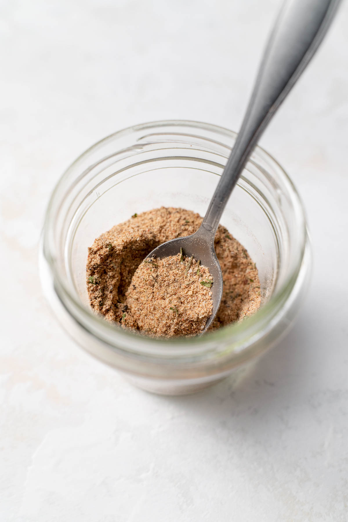 Overhead shot of a glass jar containing a spice blend, with a spoon inserted into the jar and scooping some spices. 