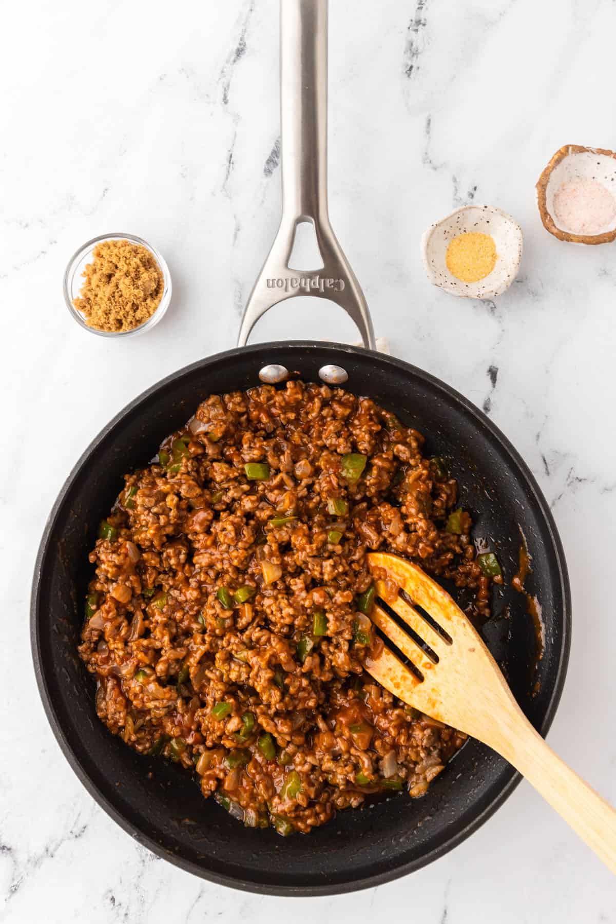 Skillet holding homemade Sloppy Joes ingredients including meat and green pepper mixture with a brown tone. A slotted wooden spoon is inserted in the mixture and there are three bowls holding spices near the top of the image.  