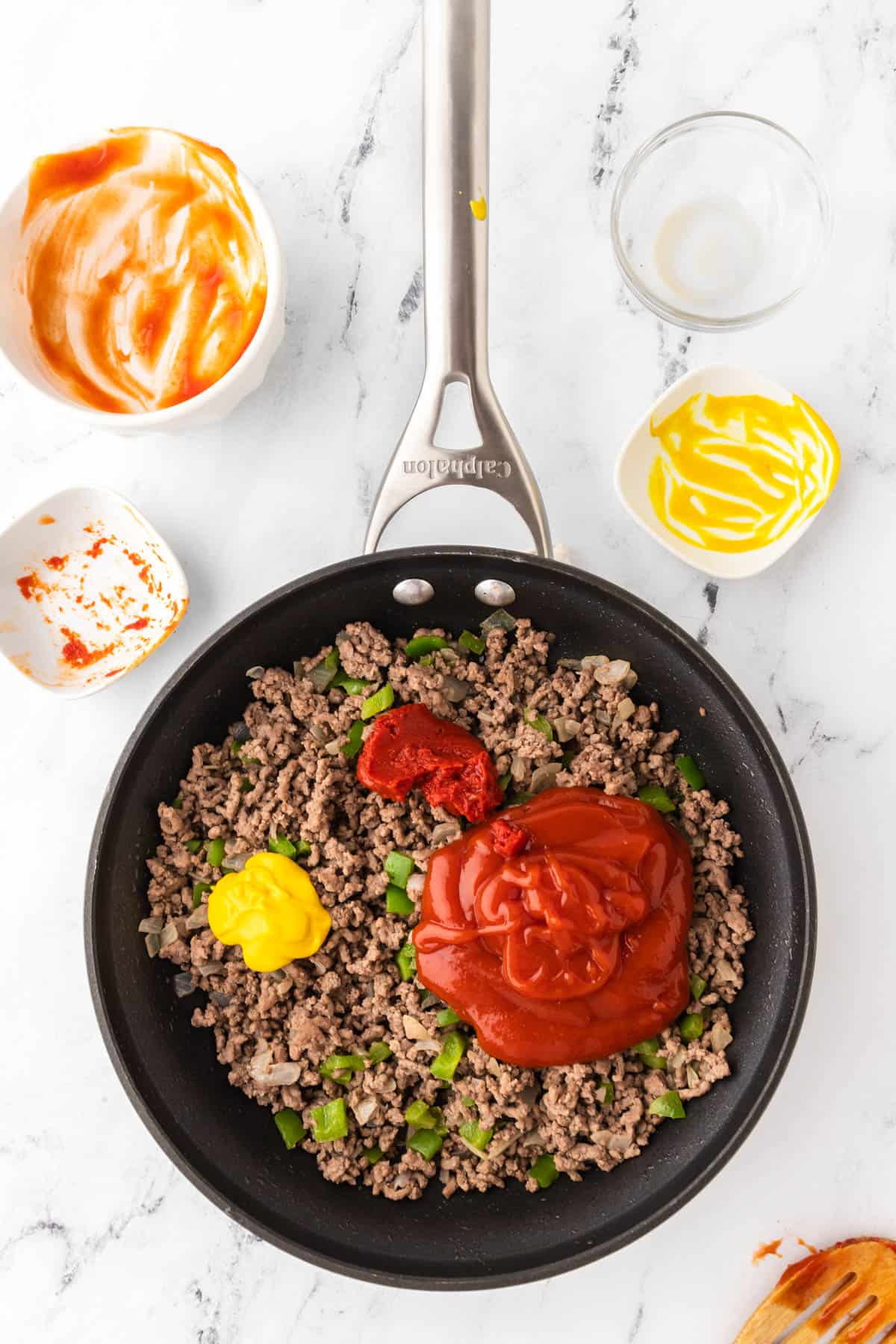 Skillet with homemade Sloppy Joes ingredients including cooked ground beef. Diced green onions are visible mixed in the meat, and ketchup, mustard and tomato sauce are visible on top of the meat mixture. Four empty bowls that have remnants of these ingredients are visible a the top of the image. 
