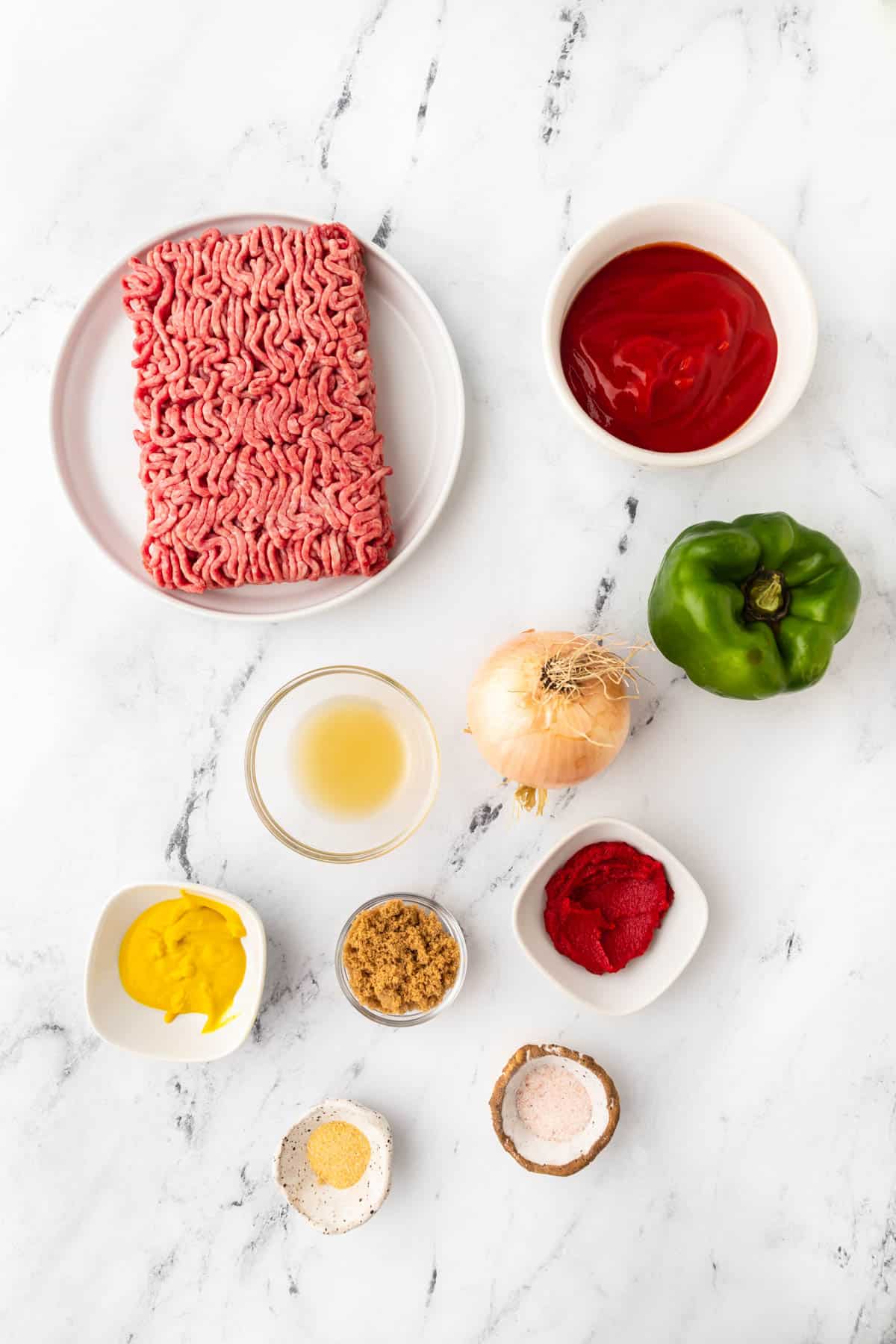 Ground beef, tomato sauce, green bell peper, onion, mustard, paprika and other spices to prepare homemade Sloppy Joes are displayed in individual bowls on a white surface. 