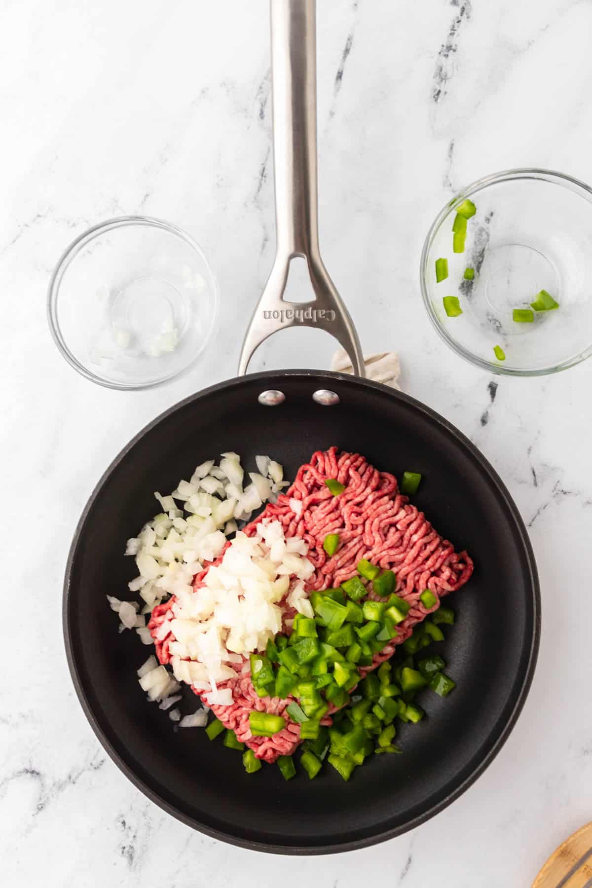 A skillet holding ground beef with finely diced onions and diced green bell peppers on top to create homemade Sloppy Joes. Empty glass bowls are displayed above the skillet. 