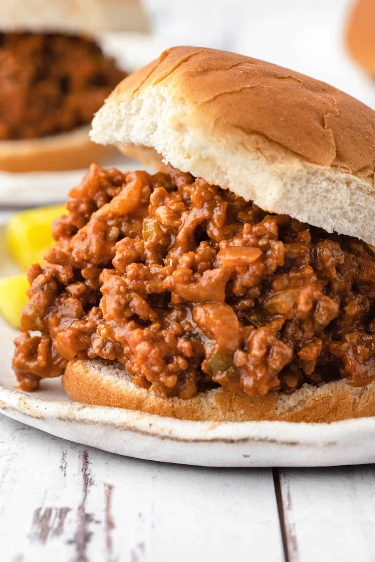Close up of homemade Sloppy Joes meat sandwiched between two hamburger buns. Pickle spears are visible in the background. 