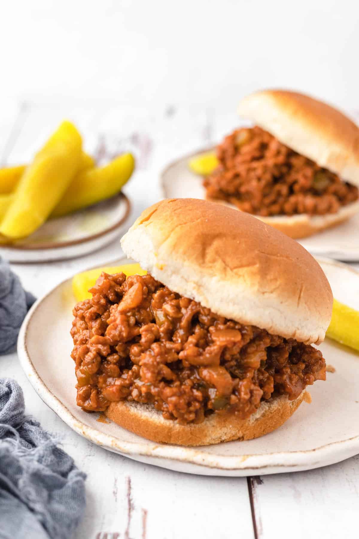 Seasoned homemade Sloppy Joes hamburger meat between two buns in the foreground. A side of pickles and a second Sloppy Joes burger in the background. 