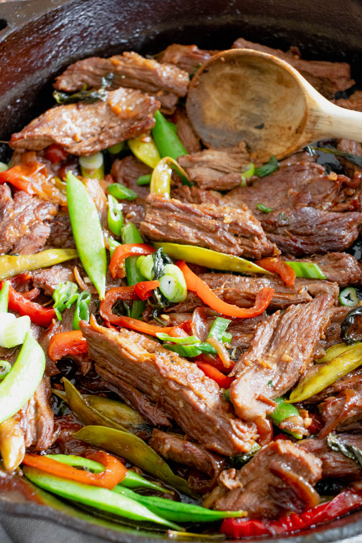 Close up image of beef and vegetable stir fry in a pan, with a wooden spoon visible near the top of the image. 