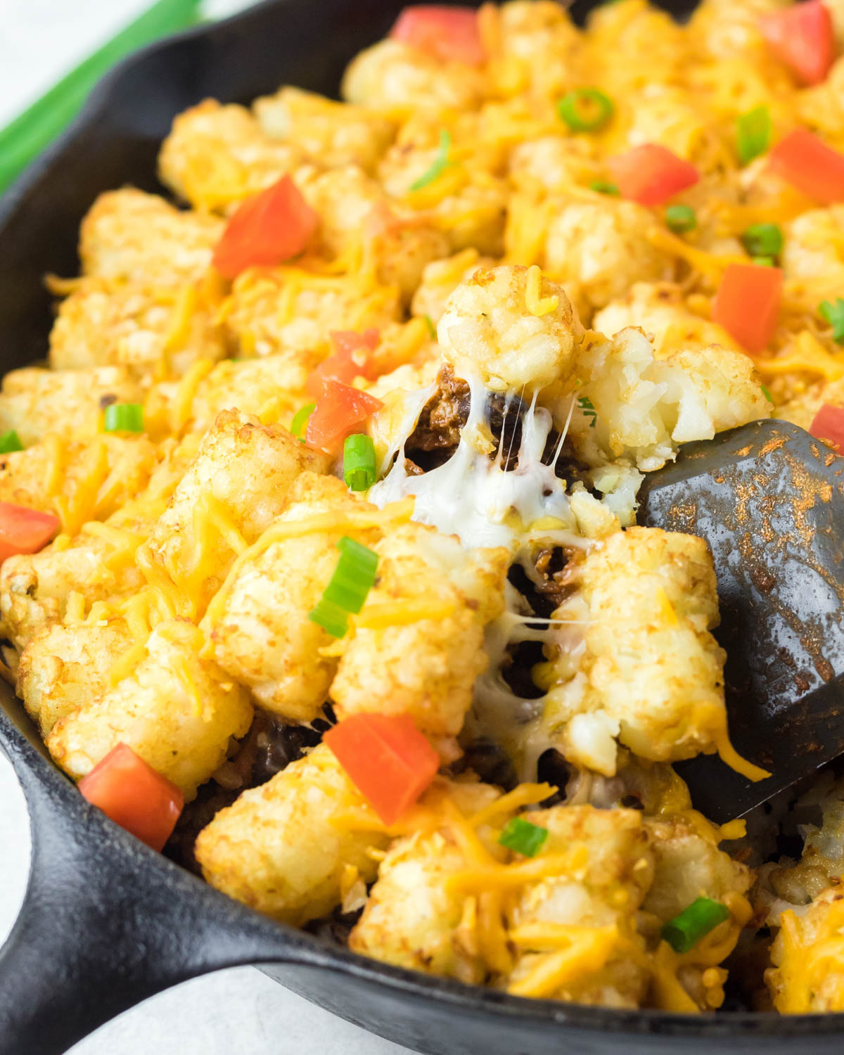 A close up shot of tater tots, melted cheese, diced tomatoes and green onions, with a spoon in a black skillet. 