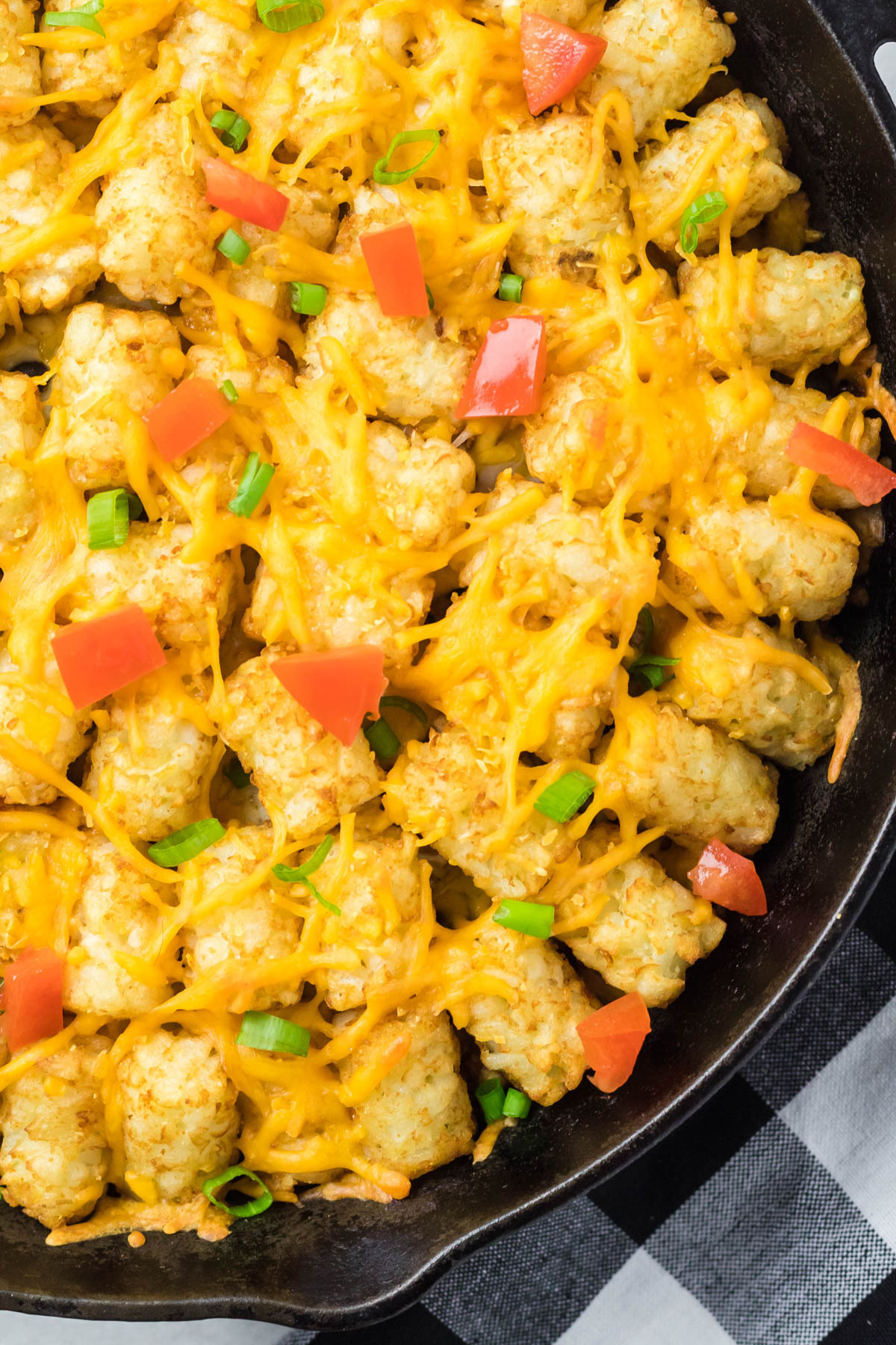 Close up image of tater tots with melted cheddar cheese and diced tomatoes and green onions in a black skillet. 