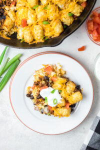 Tex Mex tater tot casserole is displayed on a white plate. Skillet full of casserole is visible at top of image, along with green onions and diced tomatoes.