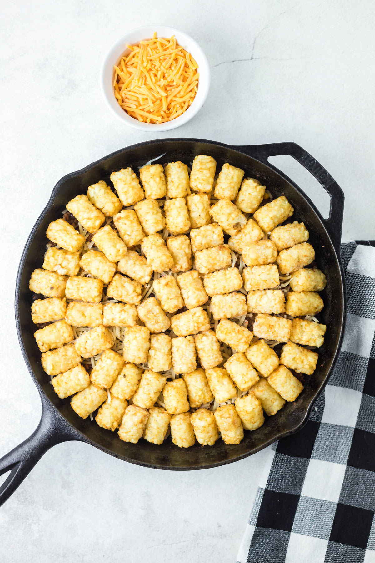 Uncooked tater tots are arranged in a single layer, with shredded cheese visible below them, in a black skillet. A bowl of shredded cheddar cheese is visible above the bowl. 