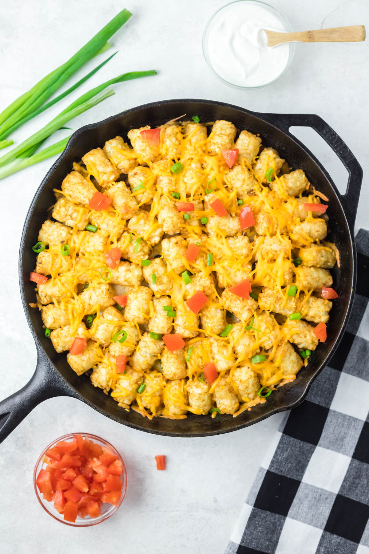 Overhead shot of black skillet containing tater tot casserole sprinkled with melted cheese, diced tomatoes and green onions. Glass bowls of extra ingredients are visible at the top and bottom of the image. 