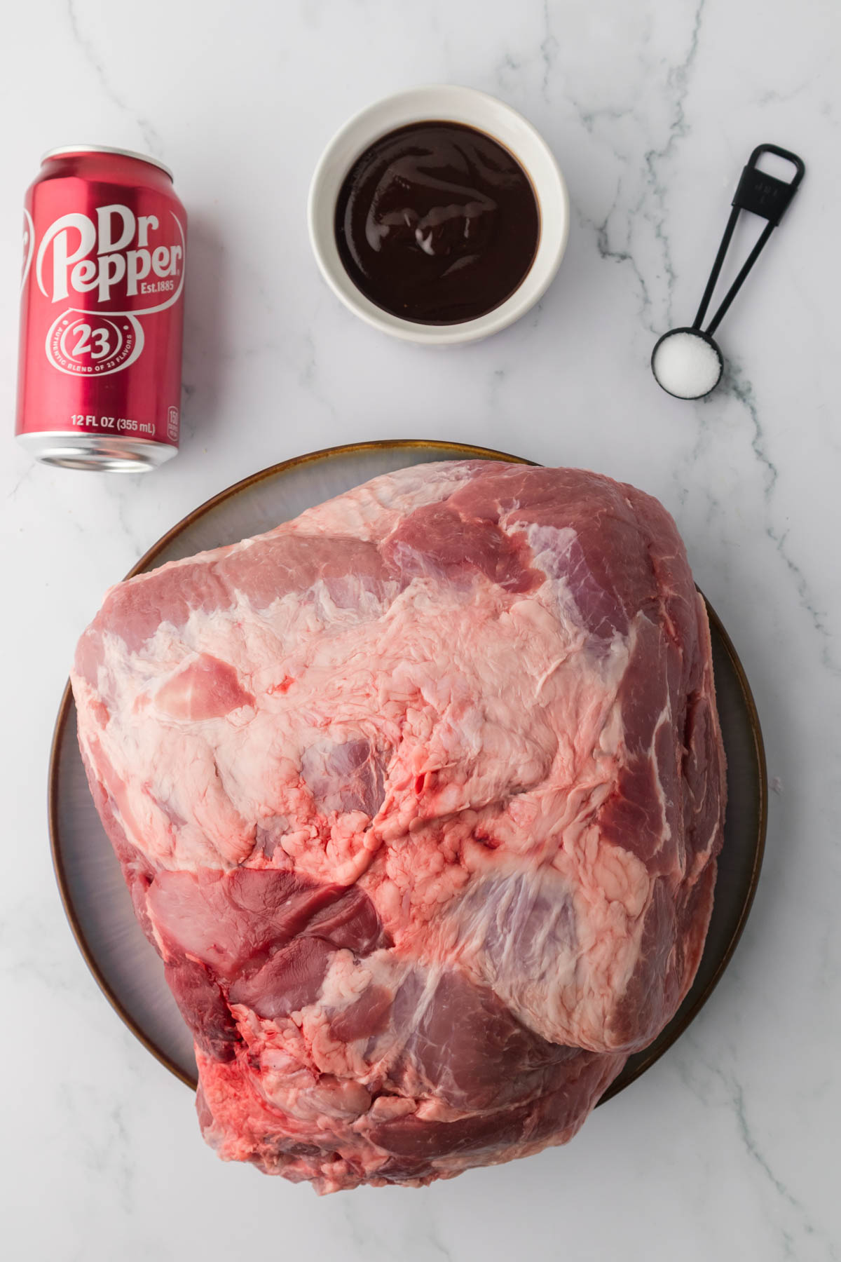 Overhead image of pork shoulder, a can of Dr Pepper, a teaspoon filled with salt, and a white bowl filled with bbq sauce. 