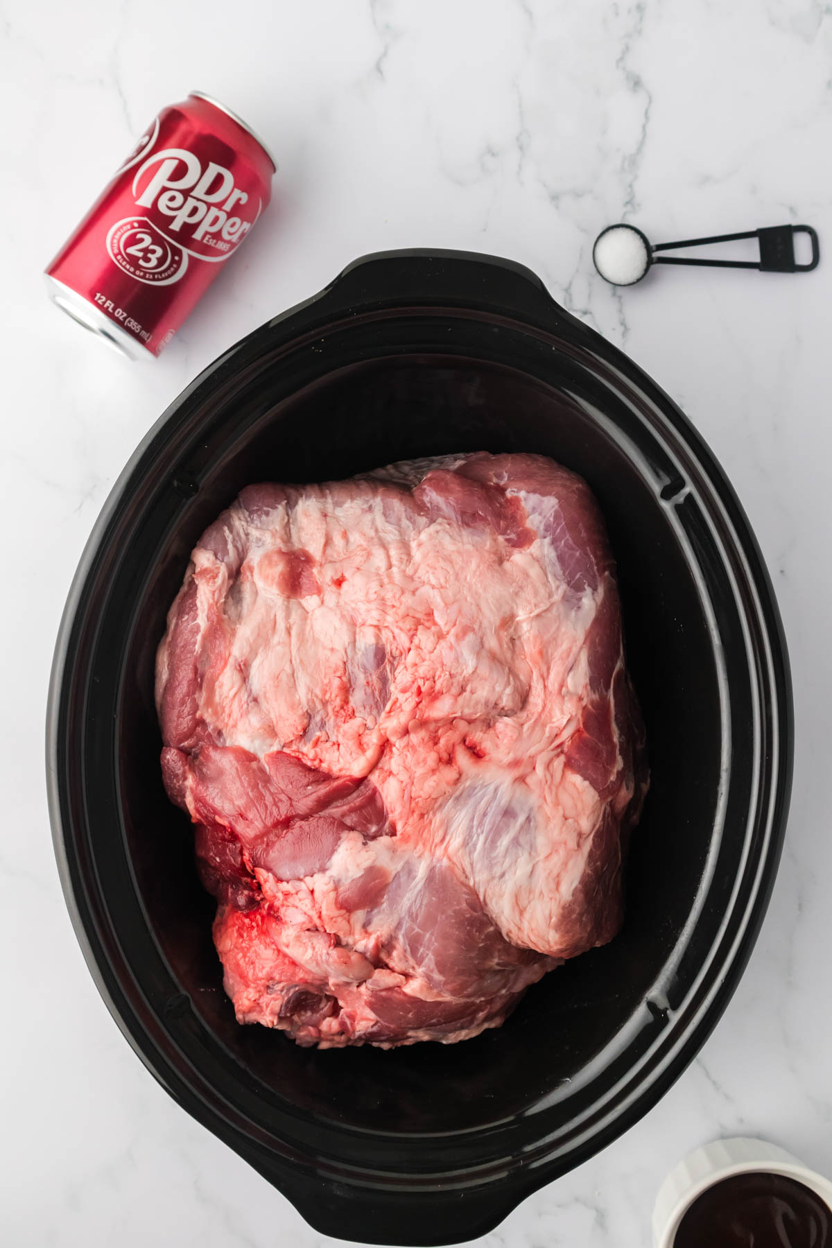 Overhead image of an uncooked pork shoulder in a slow cooker, with a can of Dr Pepper and a measuring spoon full of salt visible at the top of the image. 