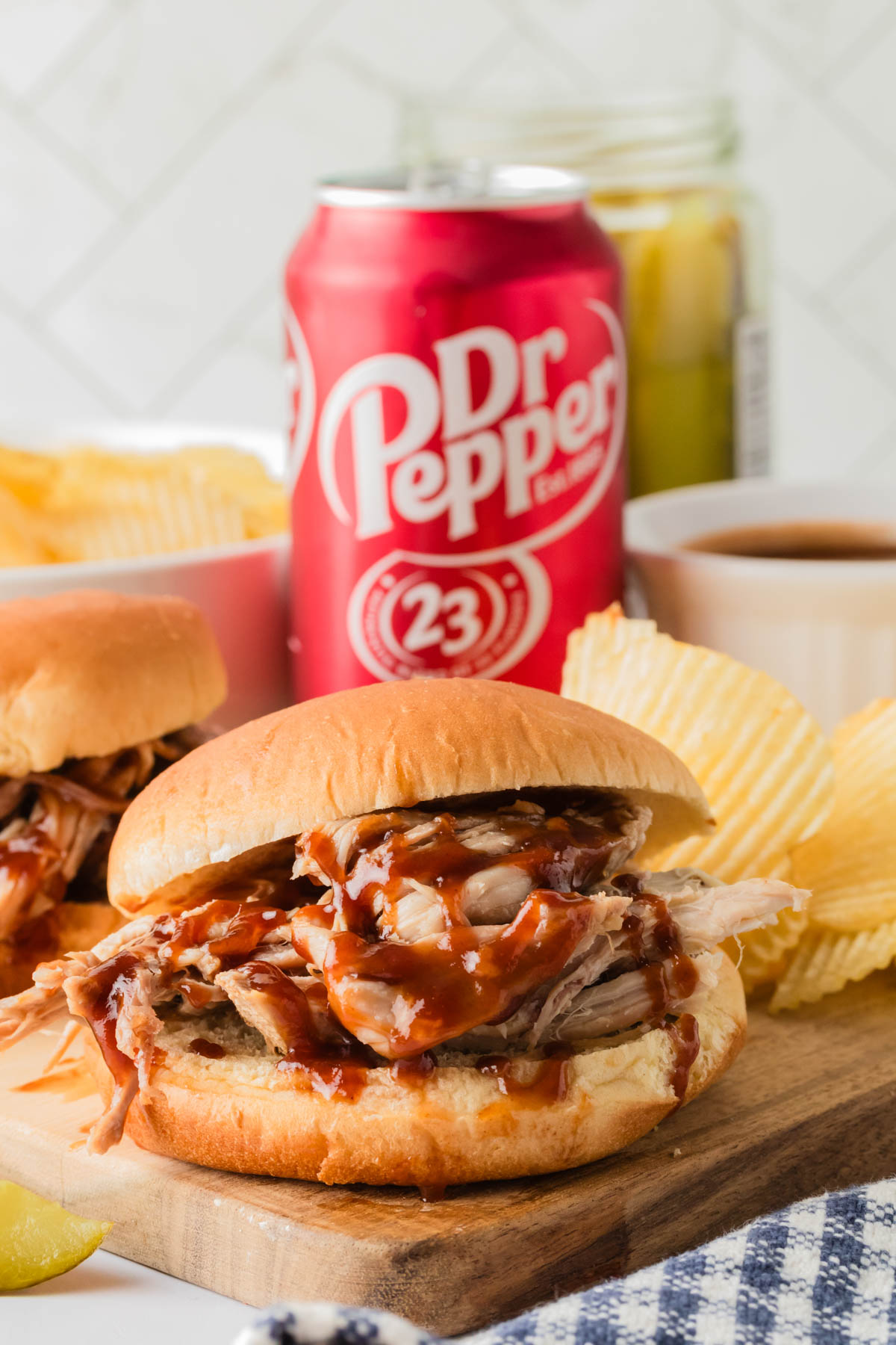 Pulled pork sandwich is featured in the foreground, drizzled with bbq sauce. Potato chips and a can of Dr Pepper are visible in the background, along with another sandwich and a bowl of sauce. 