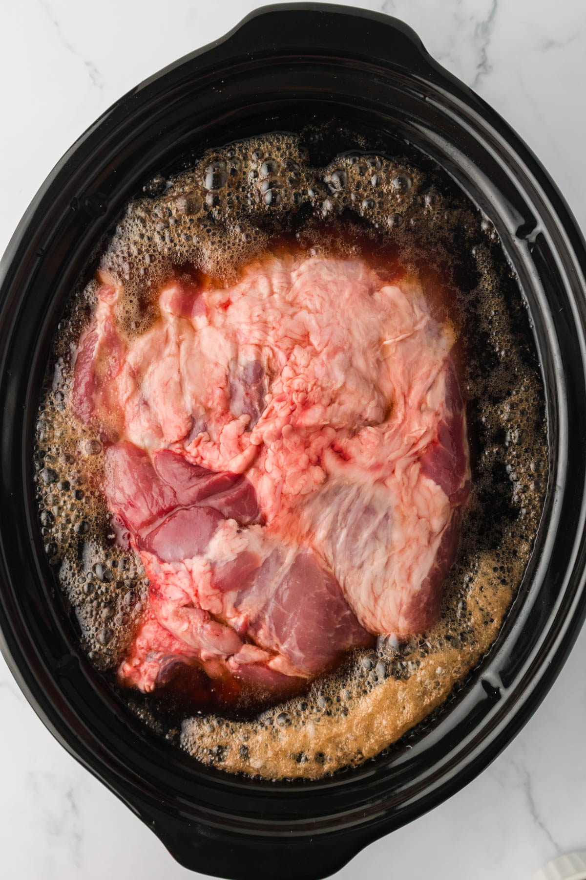 Overhead image of a slow cooker holding a pork shoulder and liquid Dr Pepper surrounding the pork. 