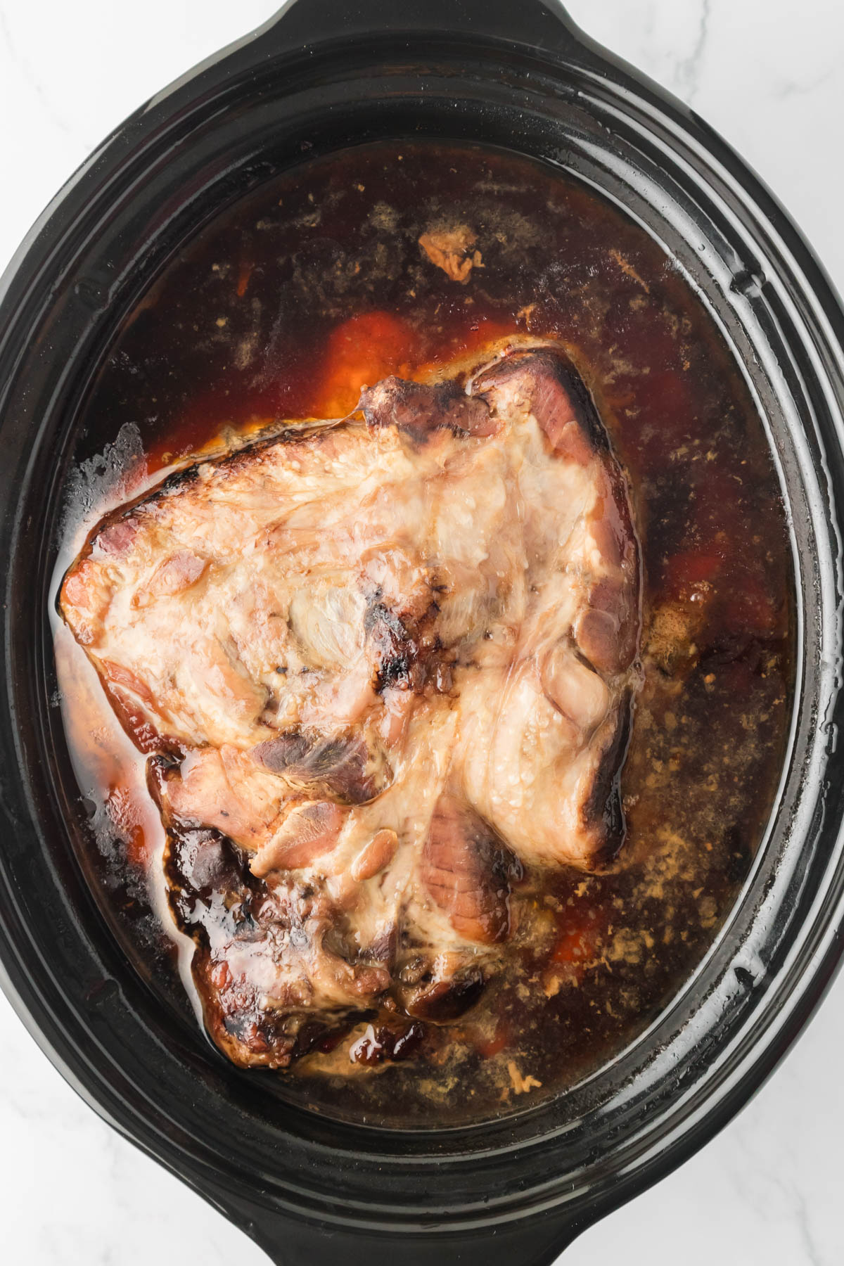 Image of pork shoulder after cooking in a slow cooker, with liquid visible around the pork. 