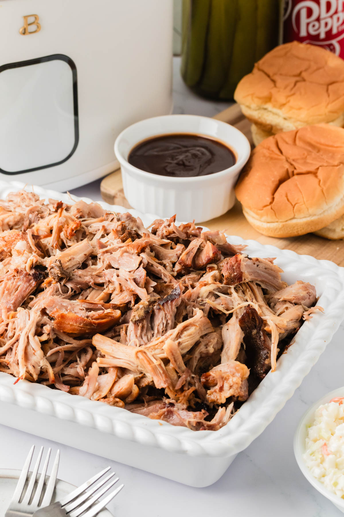 Cooked and shredded pulled pork is displayed in a tray in the foreground, with a dish of bbq sauce and hamburger buns visible in the background. 