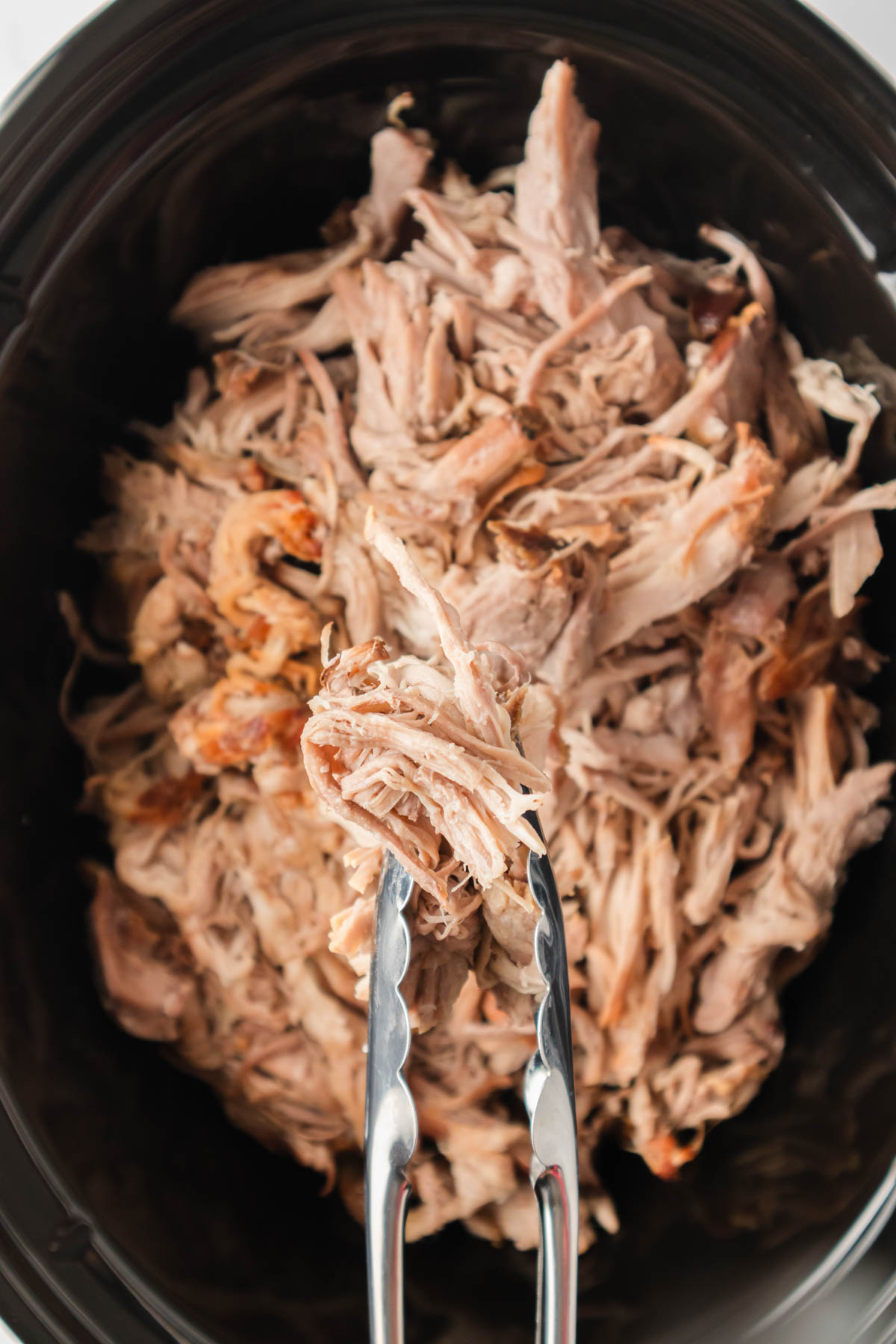 Close up image of shredded pulled pork in a slow cooker, with silver tongs visible a the bottom of the image. 
