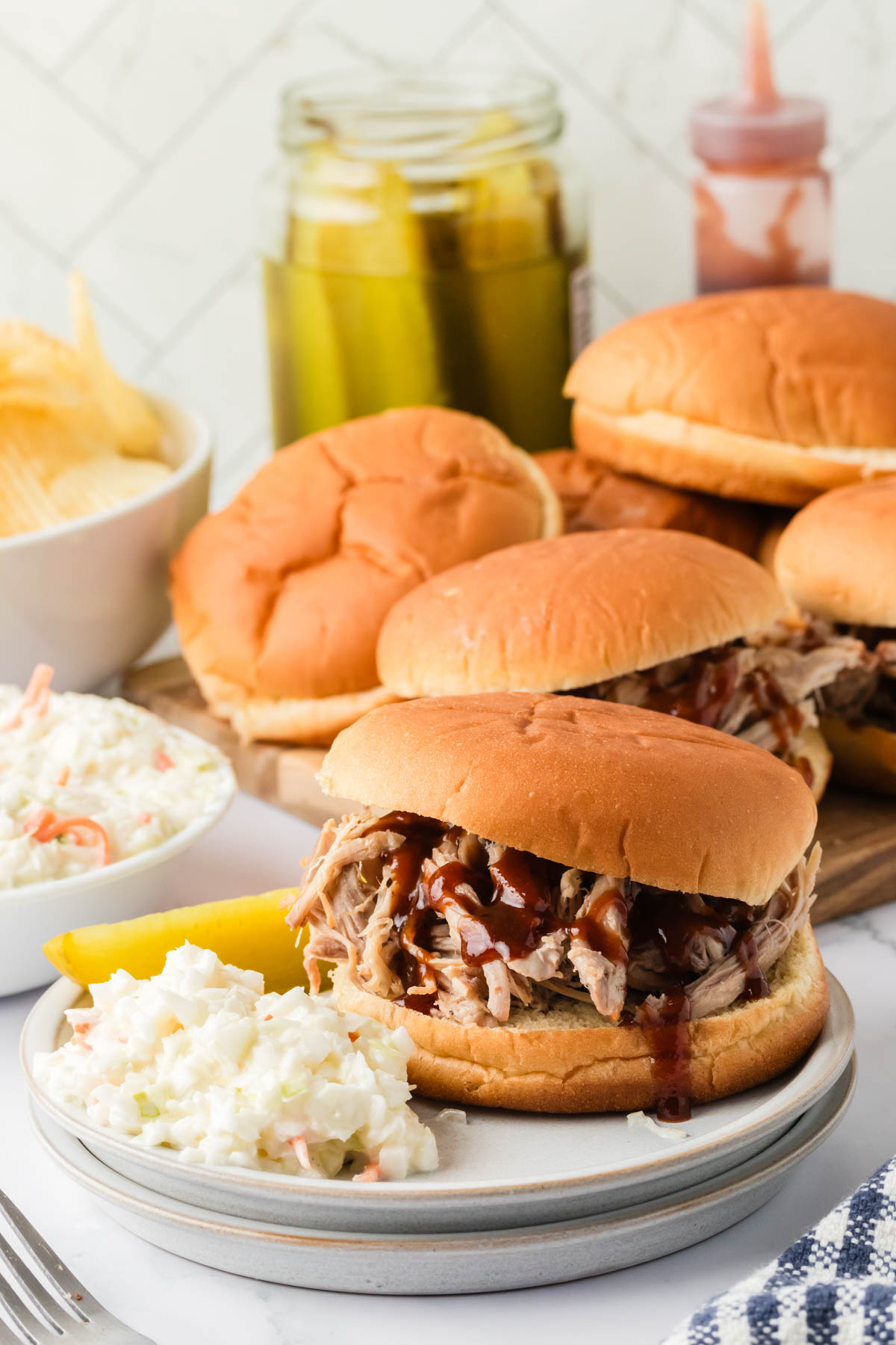 Pulled pork sandwiches are displayed on a wooden board and on a plate, with a single sandwich plated with cole slaw and a pickle spear in the foreground. 