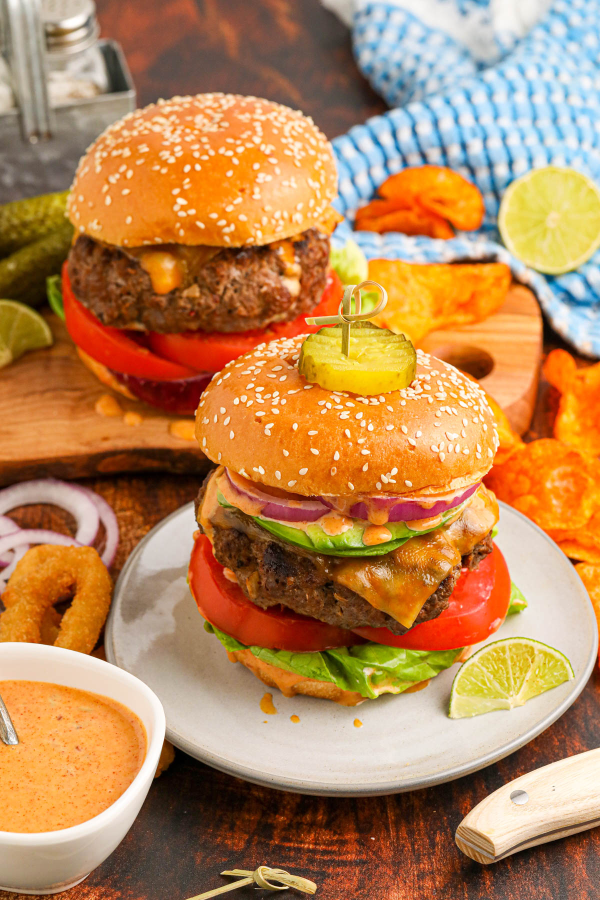 Overhead image of two hamburgers on a plate and on a wooden cutting board. Onion rings, potato chips and chipotle sauce are visible around the hamburgers.