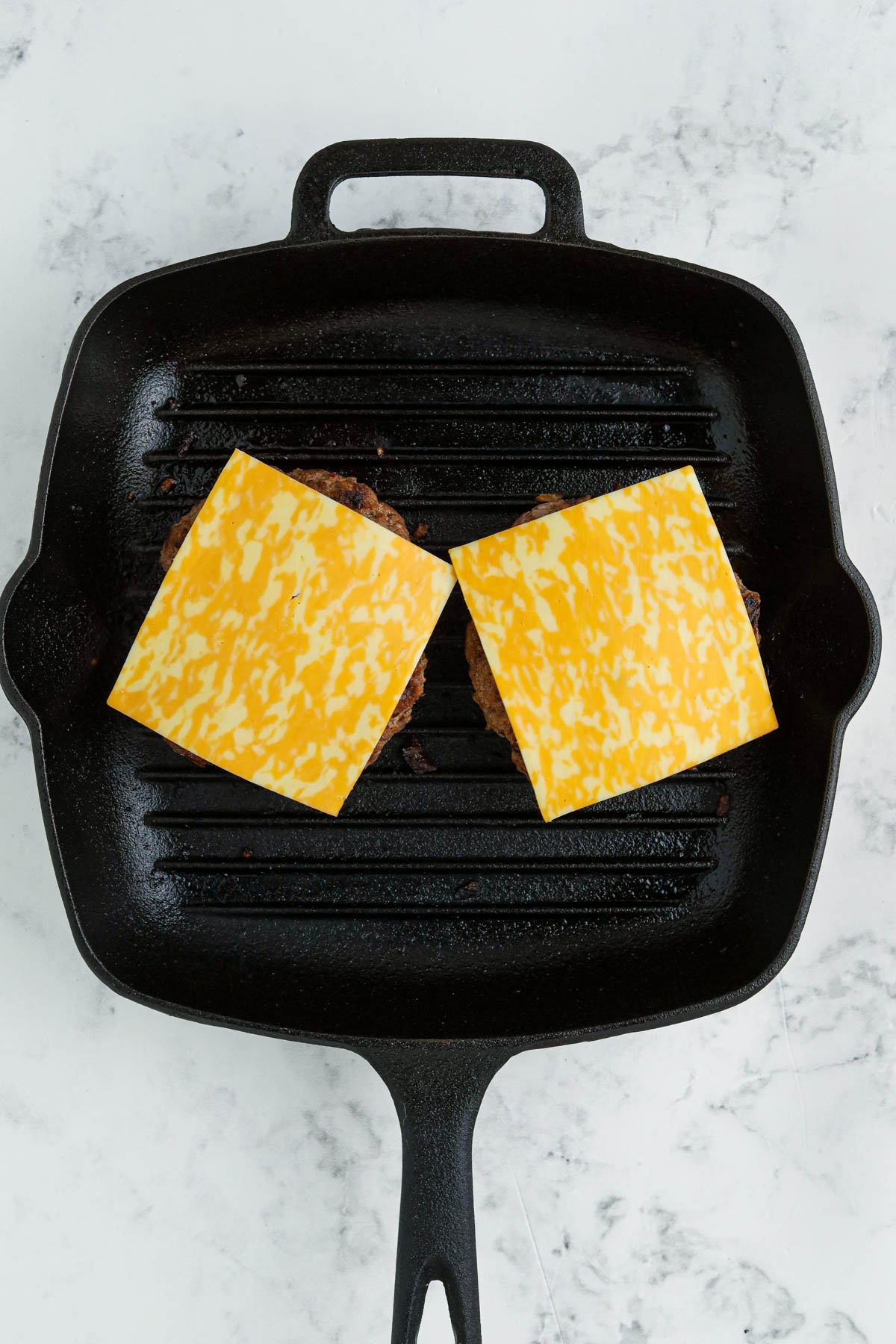 Overhead shot of a black skillet with two hamburgers topped with slices of cheese. 