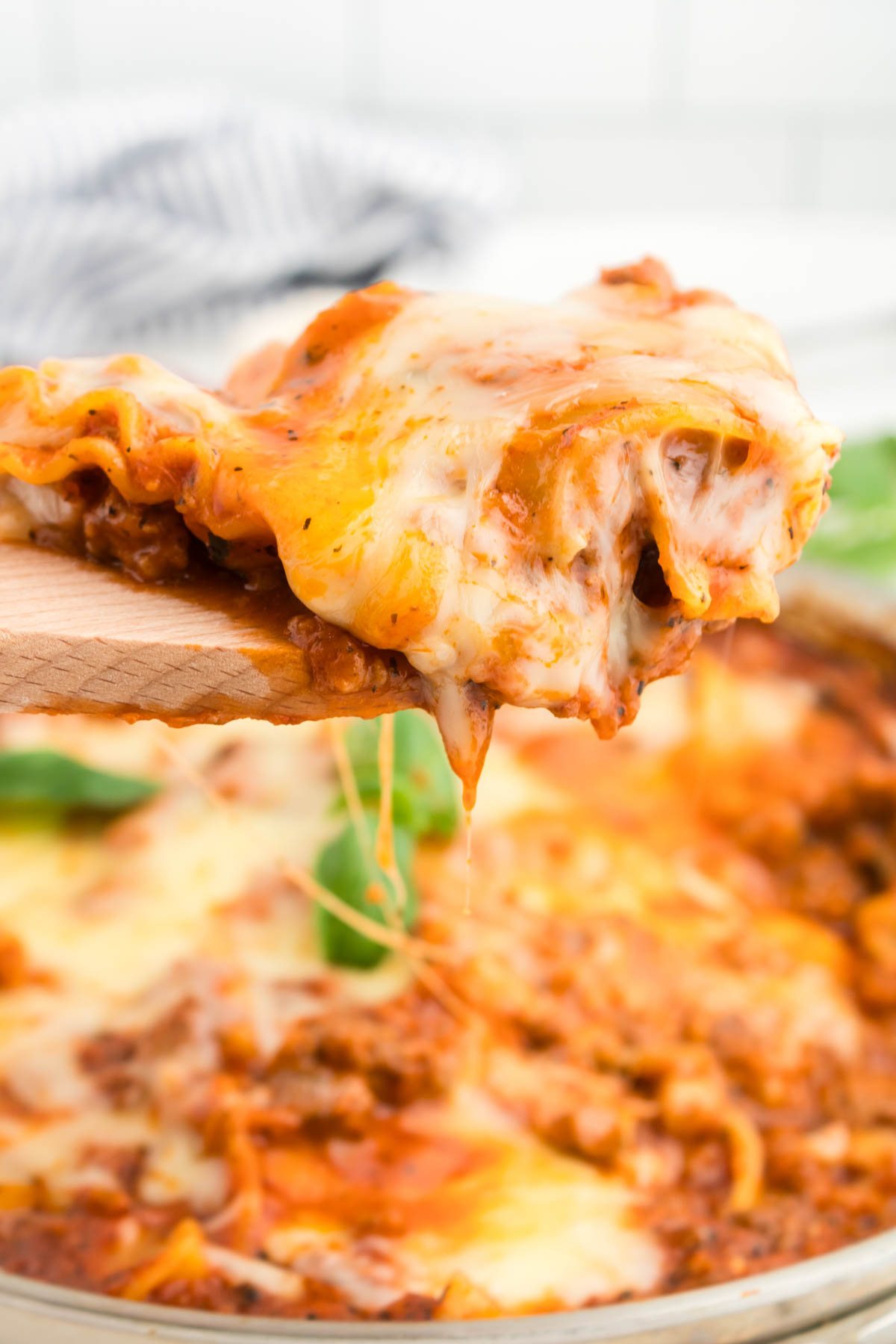 Close up shot of skillet lasagna on a wooden spoon, with a pan of lasagna visible in the background. 
