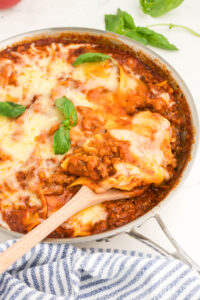 Close up image of a skillet containing cooked lasagna, with a wooden spatula inserted into the dish.