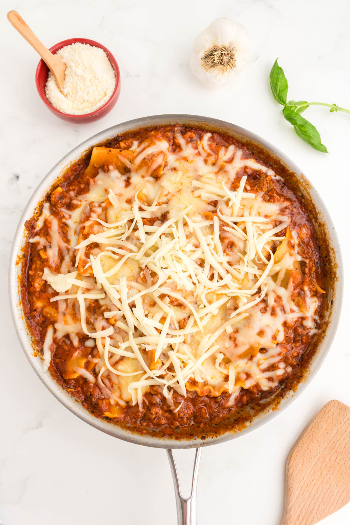An overhead view of a skillet with meat sauce and noodles, topped with cheese in various stages of melting. A bowl of parmesan cheese, a garlic clove, and a basil leaf are visible above the skillet. 