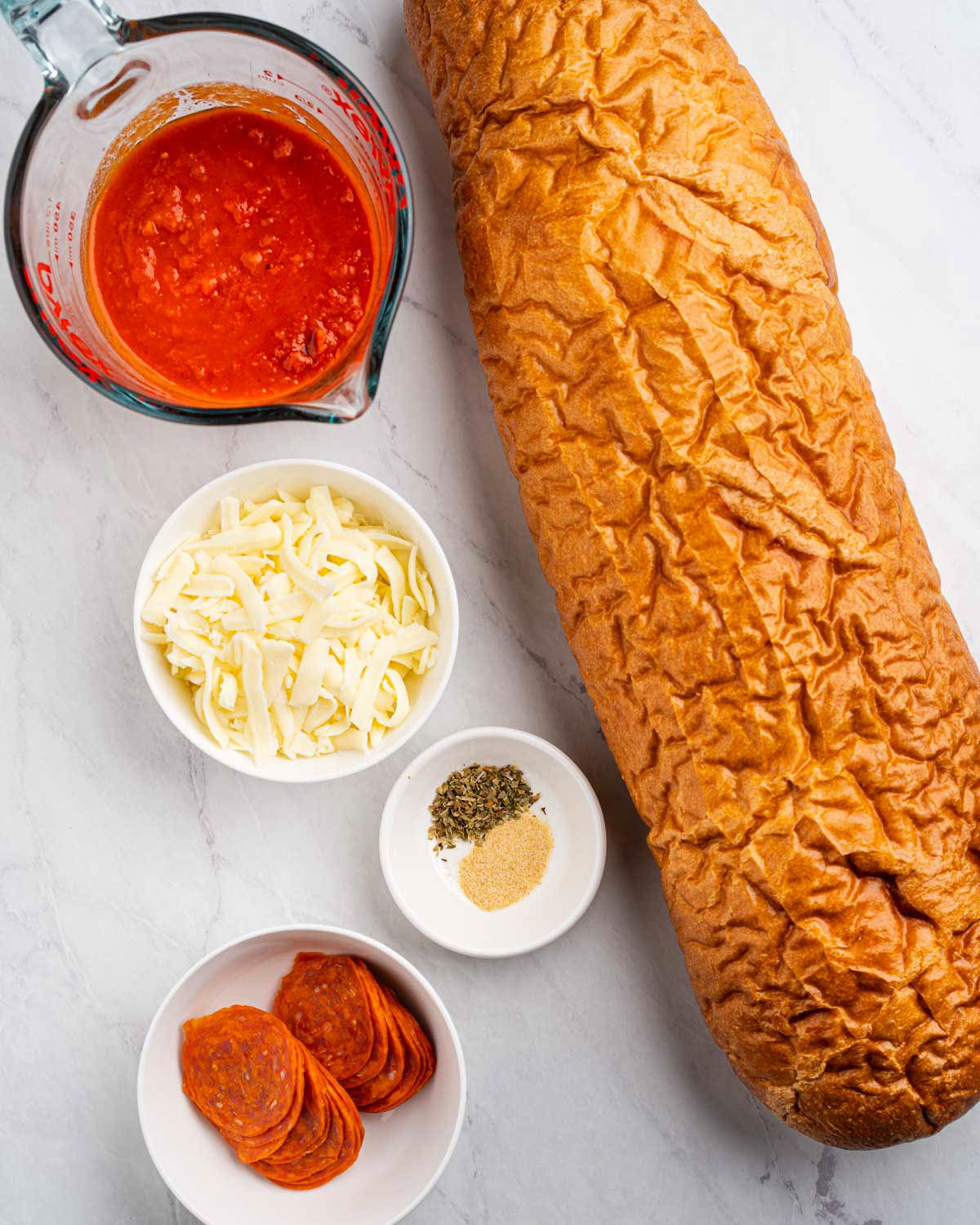 A loaf of french bread is shown, along with spices, cheese, sauce and pepperonis in individual bowls.