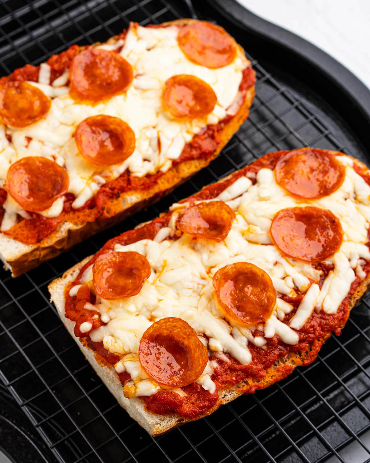 Close up image of two cooked French bread pizzas on a black cooling rack and tray. 