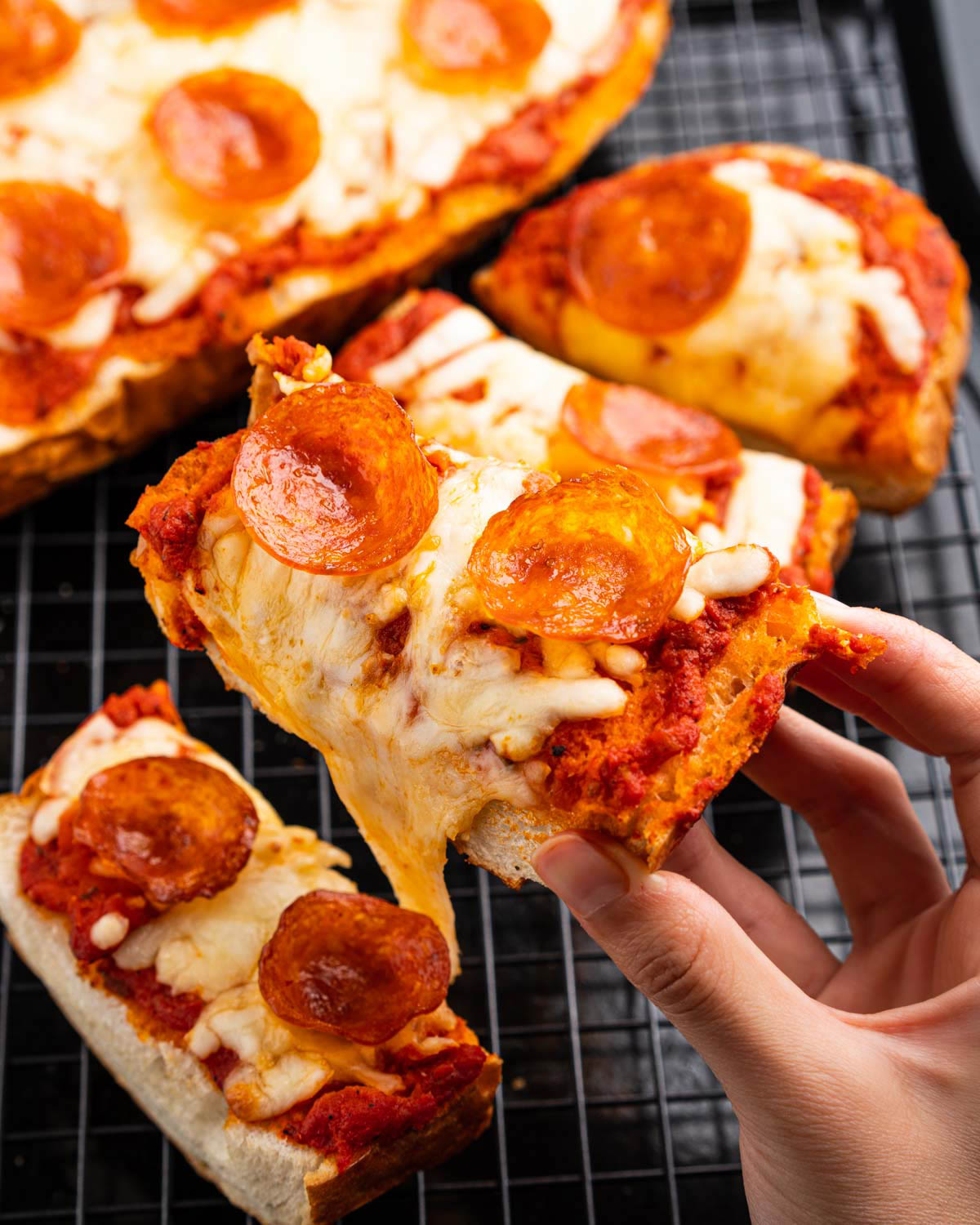Cooked french bread pizza is sliced and a hand is displaying a slice closer to the camera. 