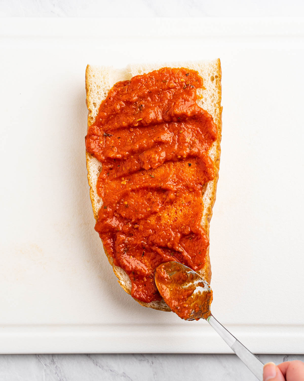Pizza sauce is being spread on a half slice of french bread. A spoon is visible at the bottom of the image. 