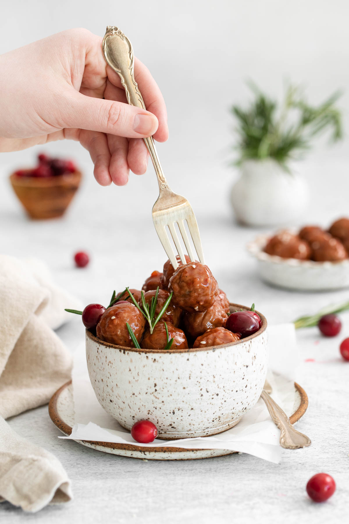 A bowl of cranberry meatballs are displayed, with a hand holding a fork in the process of selecting a meatball. Other bowls and a sprig of rosemary are visible in the background. 